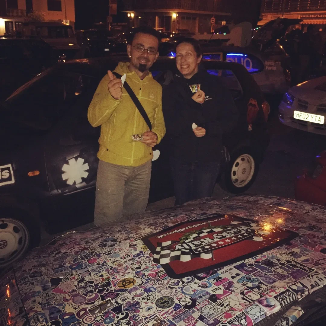Two people standing in front of a car covered with stickers at night, smiling and showing thumbs up.