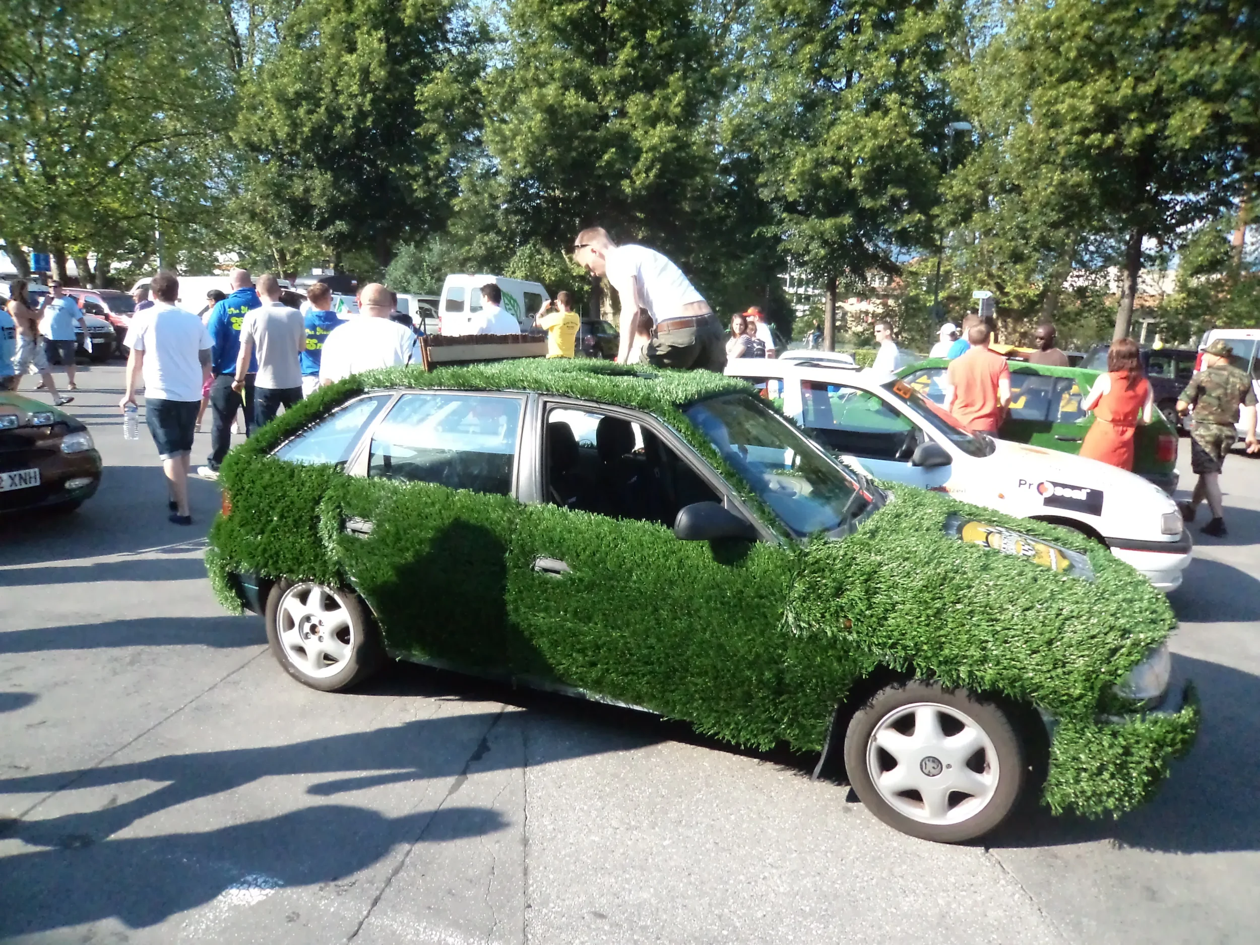 A car covered in green artificial grass at an outdoor event with dozens of people walking around in the background.