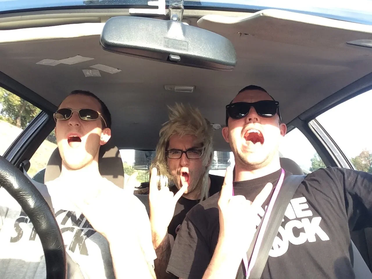 Three young men in a car making rock hand gestures, wearing sunglasses, with enthusiastic expressions, sunlight inside the vehicle.