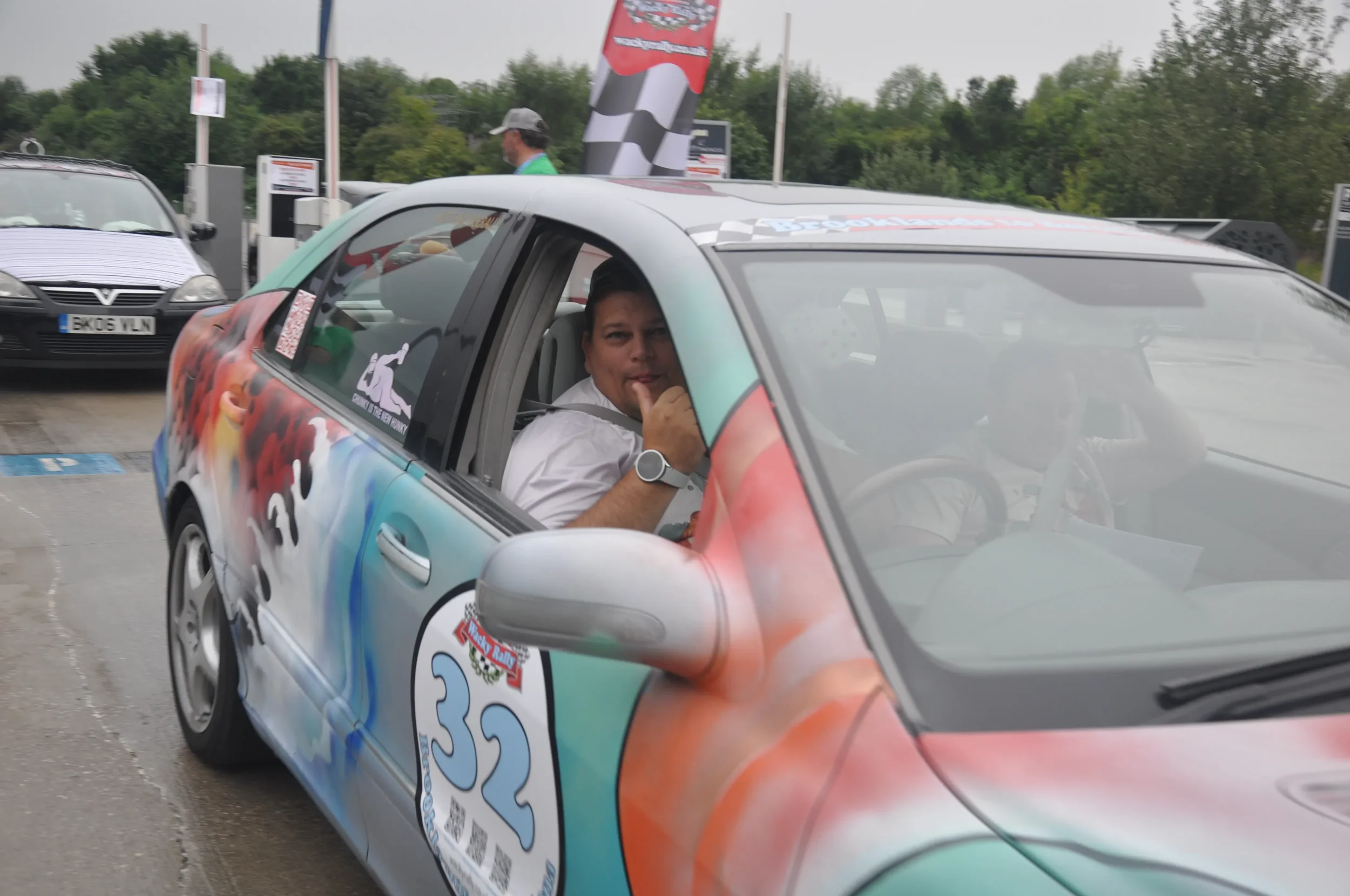A man with a watch in the driver's seat of a colorful race car, giving a thumbs up through the window, at a rally event with other cars and a flag in the background.
