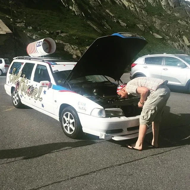 A man inspecting the open hood of a decorated white police-themed car with a large soda can on the roof, parked on the side of a mountain road, with other cars nearby.