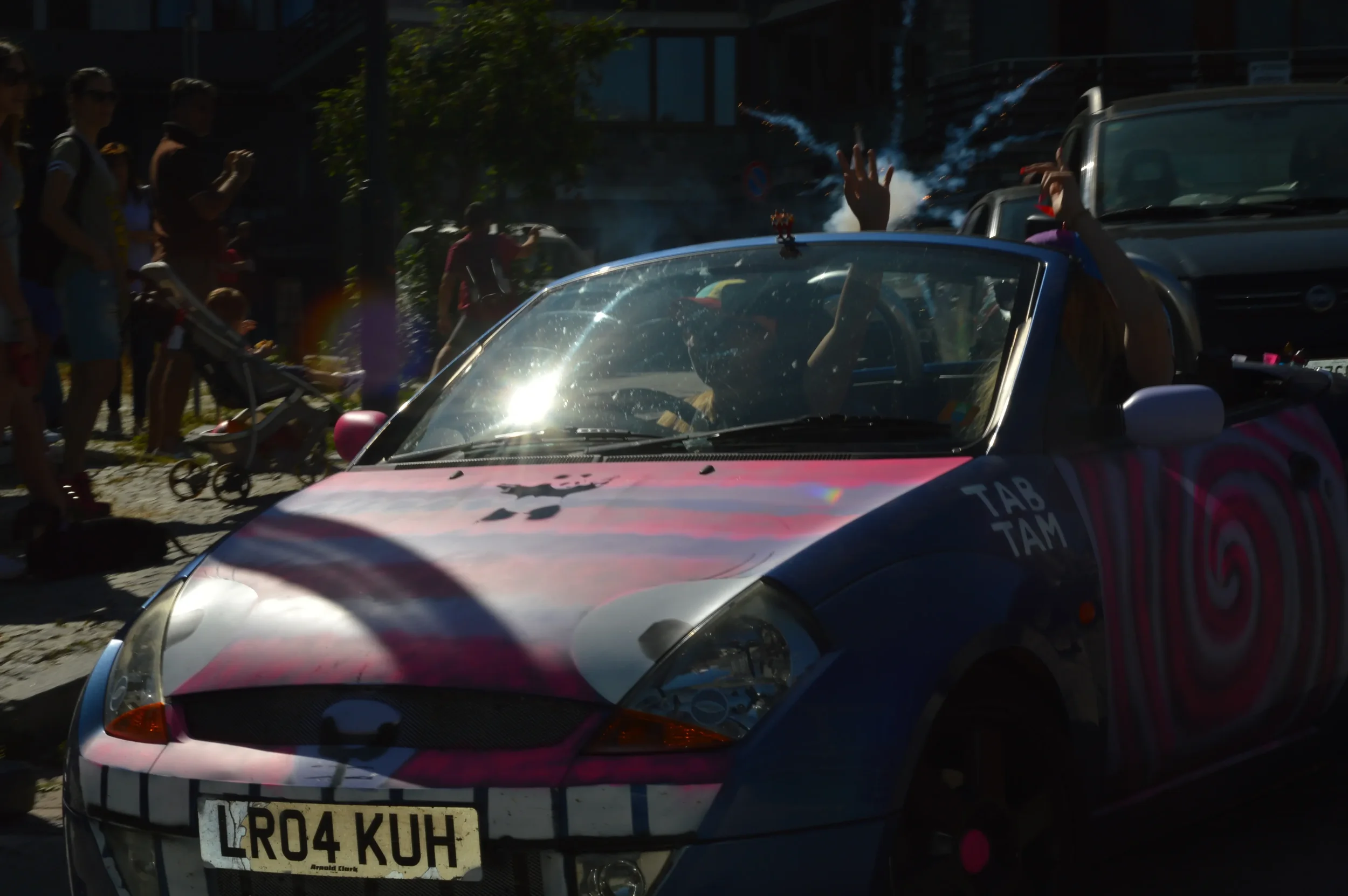 Colorful convertible car decorated with art, surrounded by people at a parade or event, with some audience members taking photos.