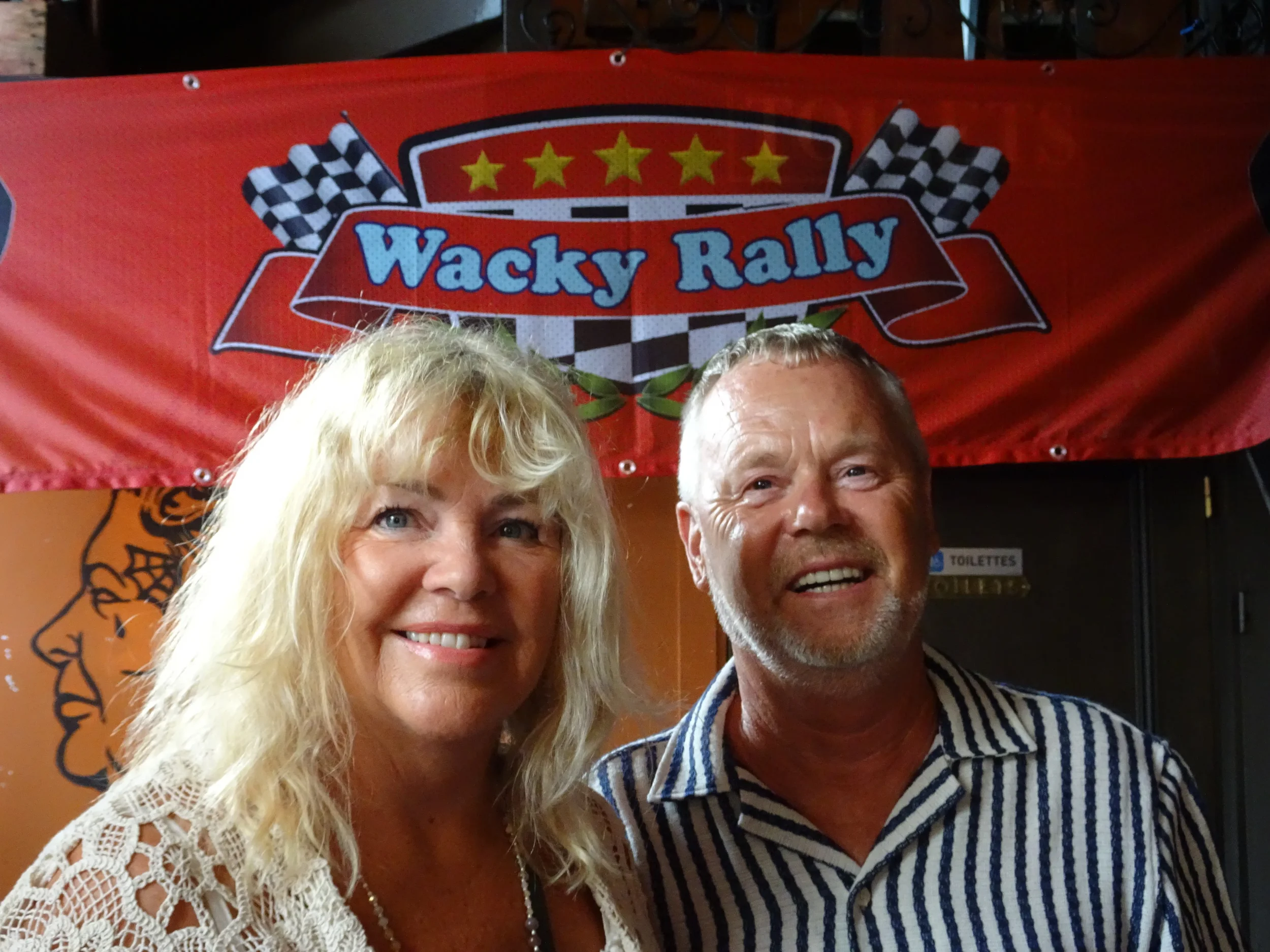 Two smiling people, a woman with long blonde hair wearing a lace top, and a man with short gray hair and beard wearing a striped shirt, standing in front of a red banner that reads 'Wacky Rally' with checkered flags.