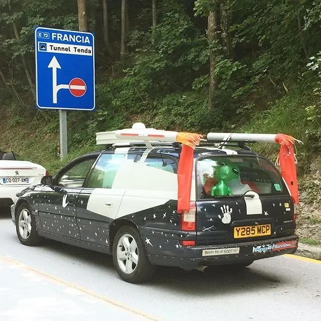 A decorated car with space-themed decals, including moon and star images, parked by the side of a road near a forest. The car has a large inflatable green alien in the back window and an orange ribbon tied around a pole on the roof. There is a blue r