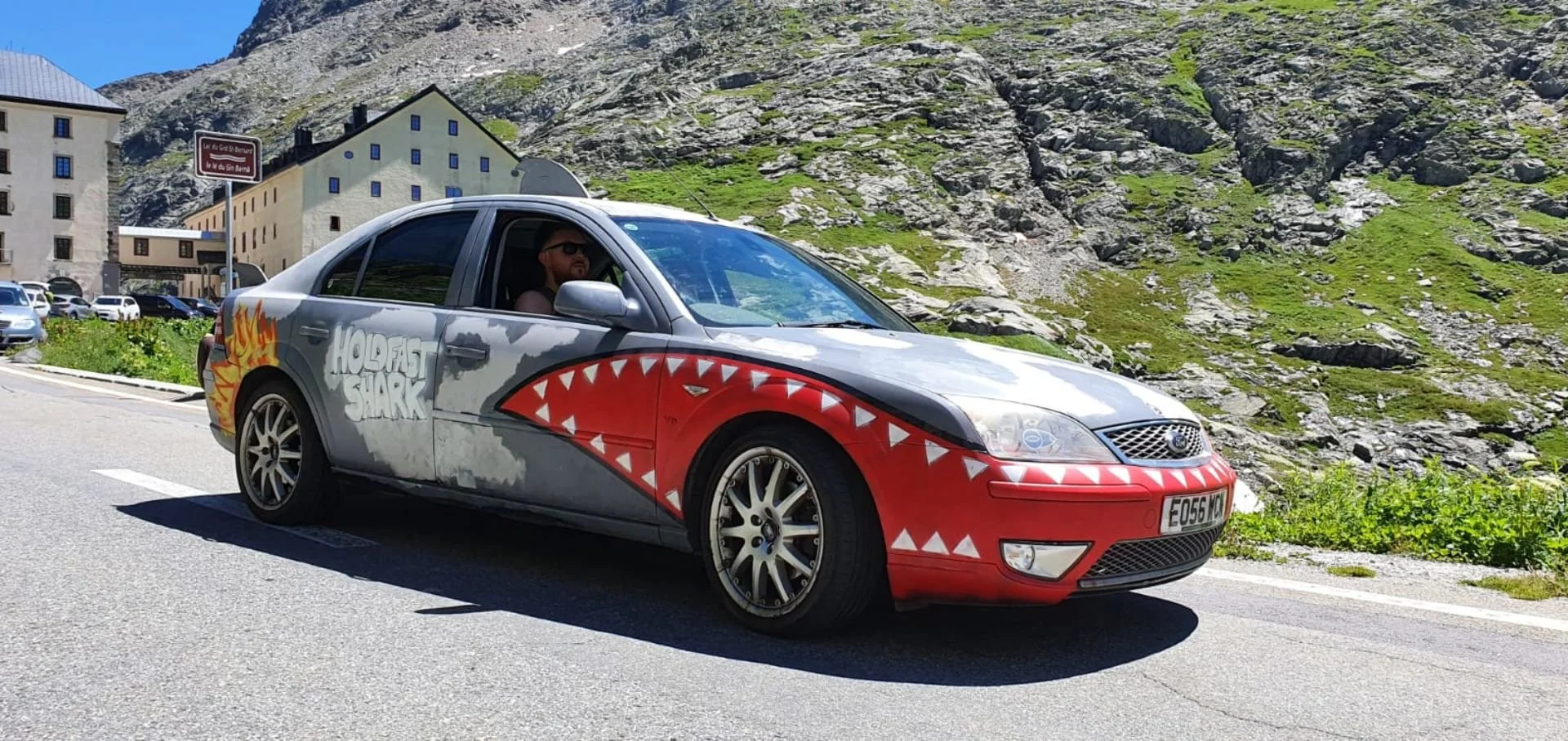 A silver and red car with parts shaped like a shark's mouth and a flame on the rear side, parked on a mountain road, with mountains and buildings in the background.