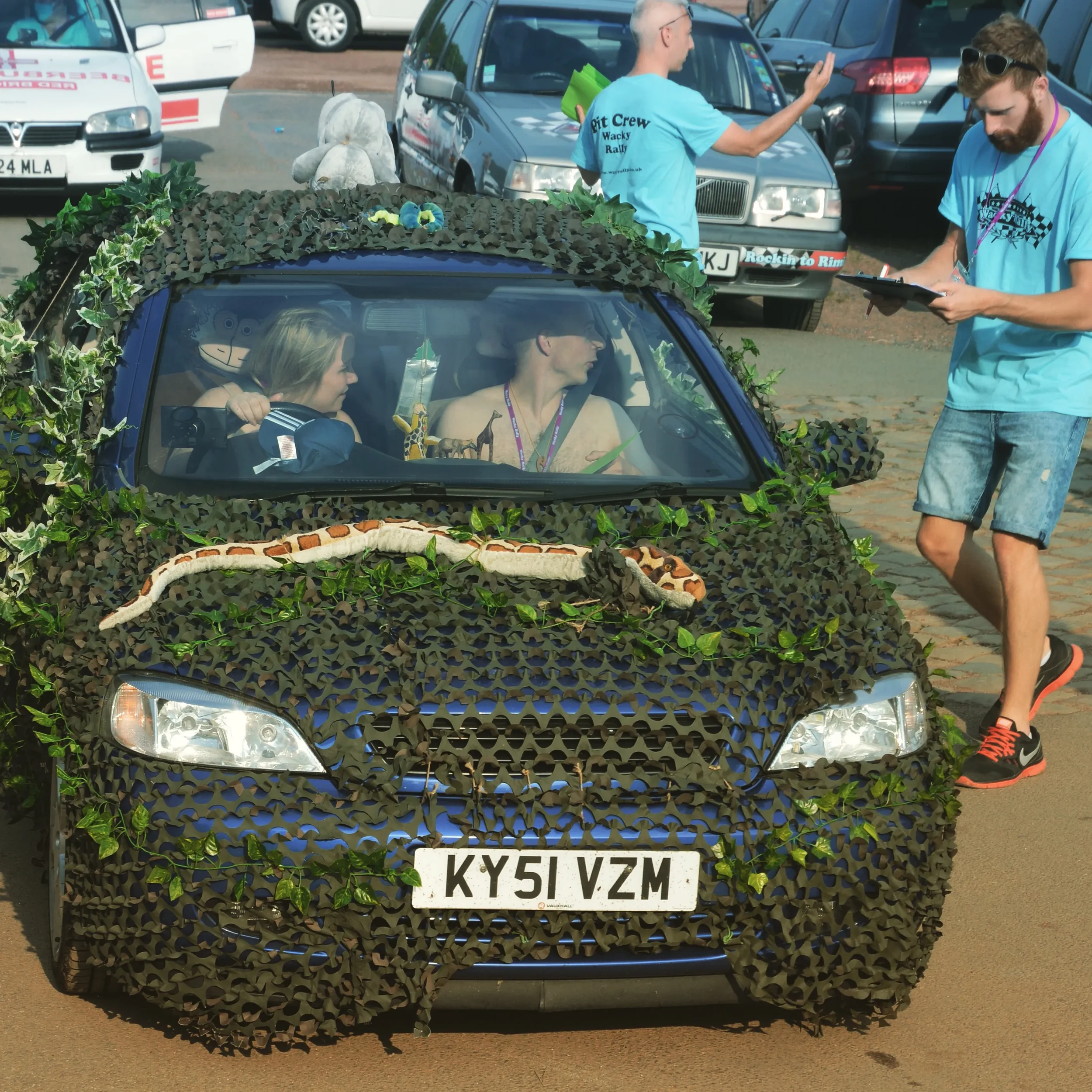 A car covered in camouflage netting with the license plate KY51 VZM, decorated with greenery and faux animals, at a rally event. Two women are inside, one driving and the other in the passenger seat, and two men in blue shirts are standing outside, o