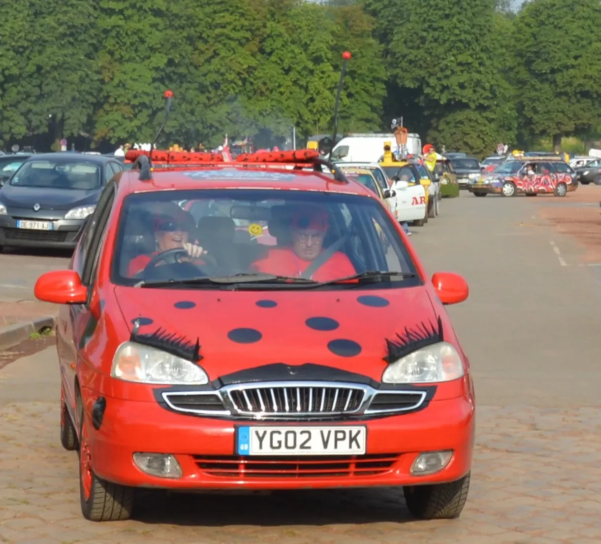 A red car with painted ladybug spots and eyelashes on the headlights, driving in a crowded parking lot with many other vehicles, trees, and people in the background.