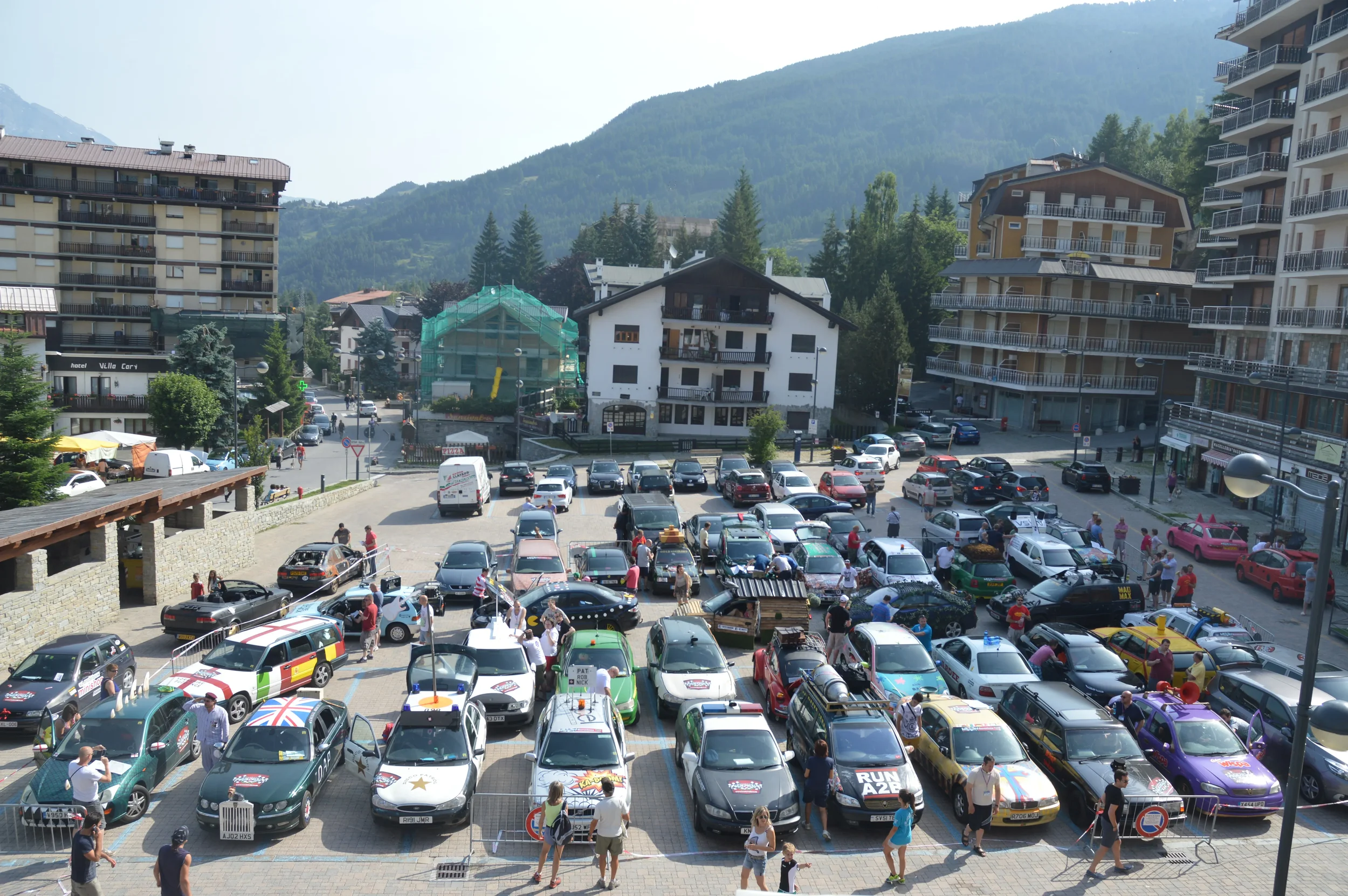 A busy parking lot filled with various colorful cars and people walking around, with buildings and mountains in the background.