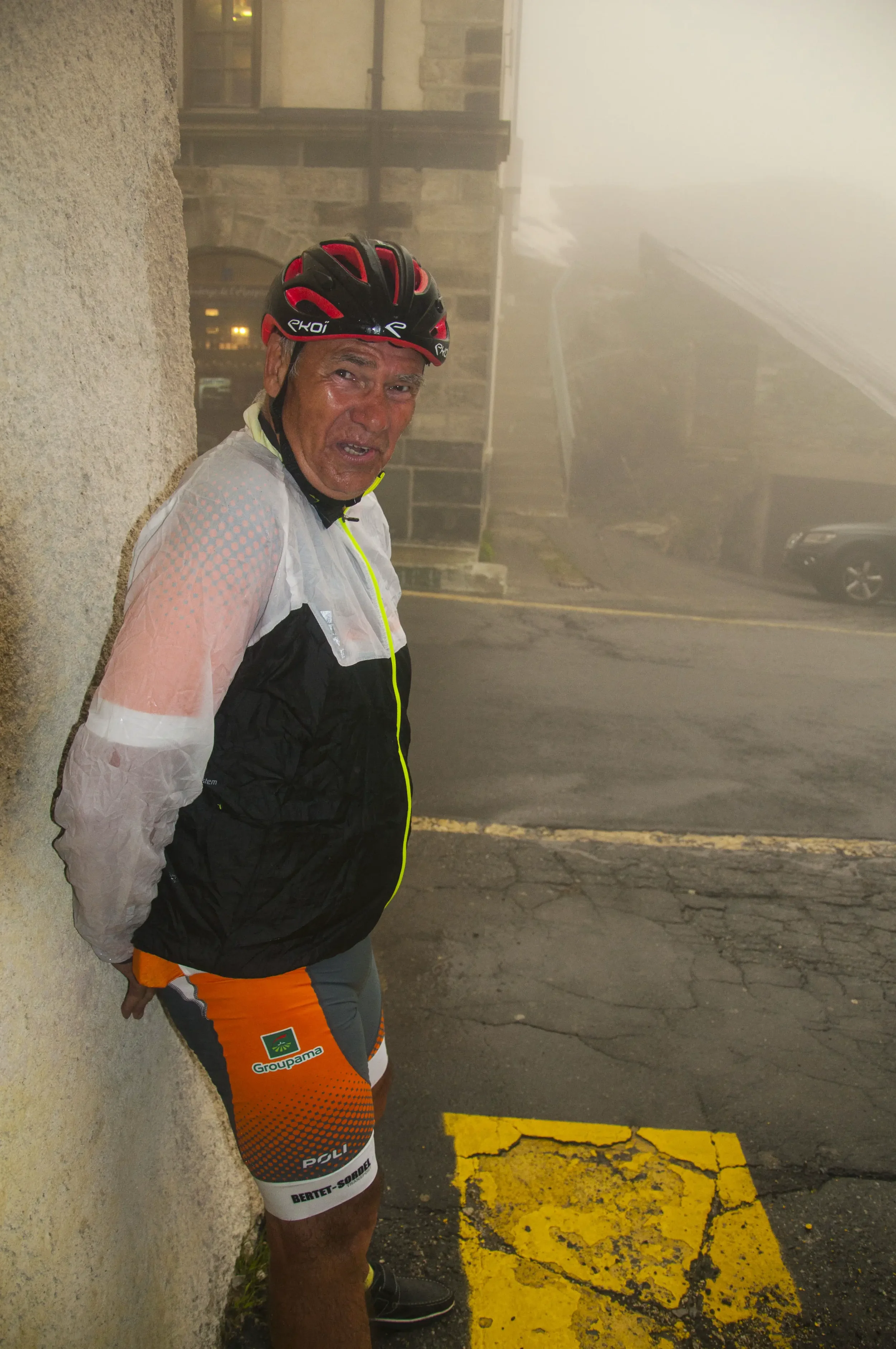 A man in cycling gear and a helmet leaning against a wall in foggy weather on a paved street.