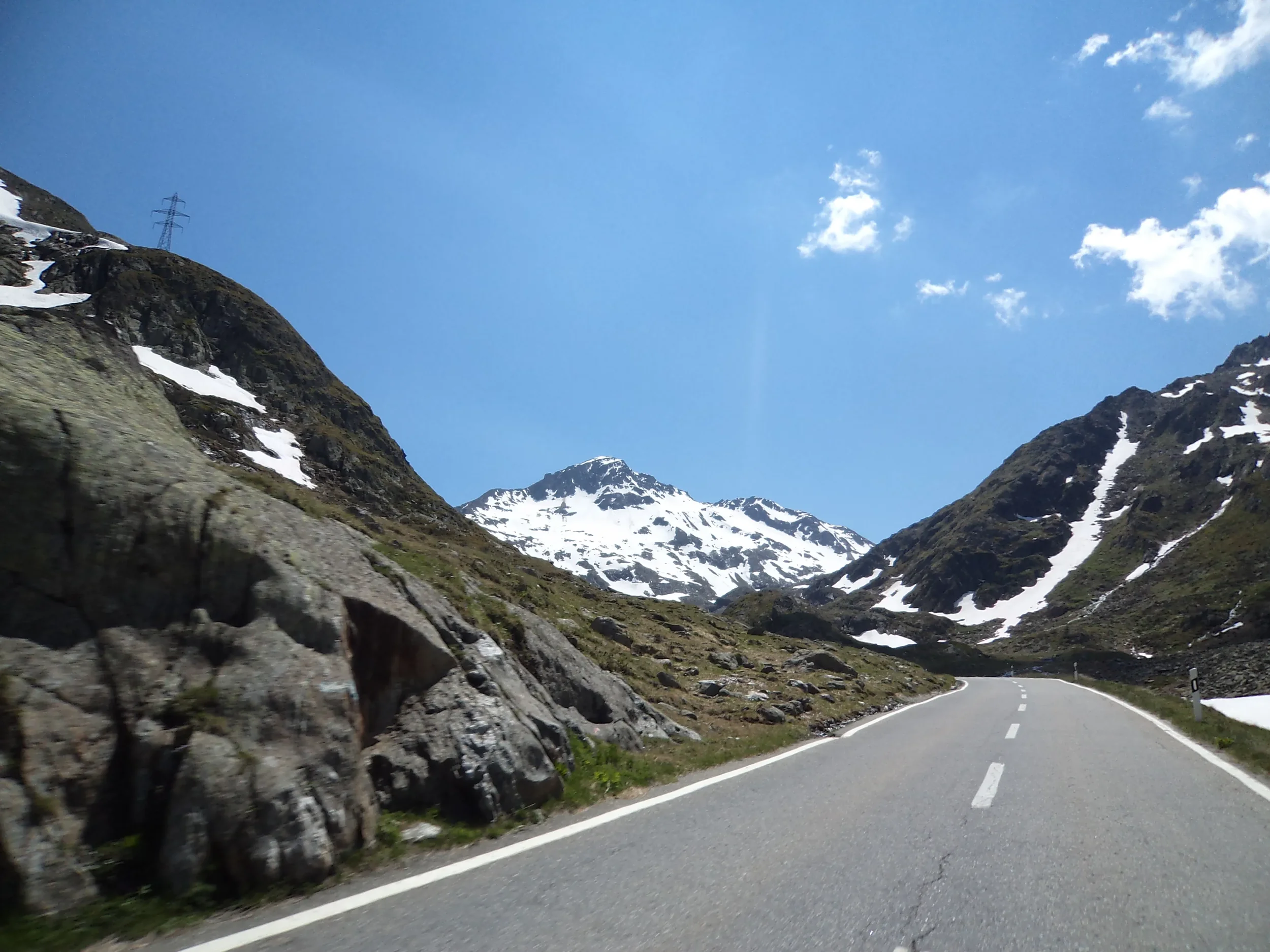 A mountain road winding through snow-capped mountains under a blue sky with some clouds.