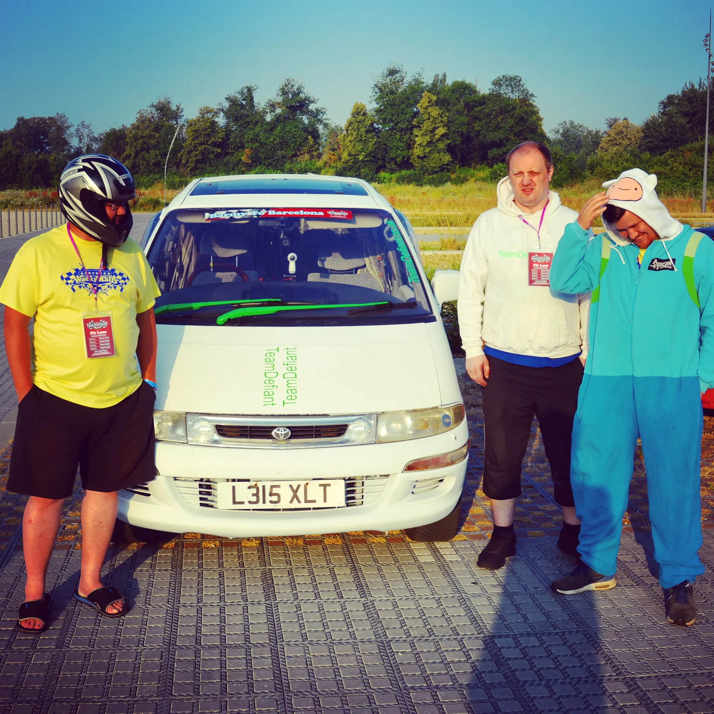 Three men standing next to a white racing car on a paved area with green trees in the background. One is wearing a helmet and yellow shirt, another is in a white hoodie, and the third in a blue onesie with a hat.