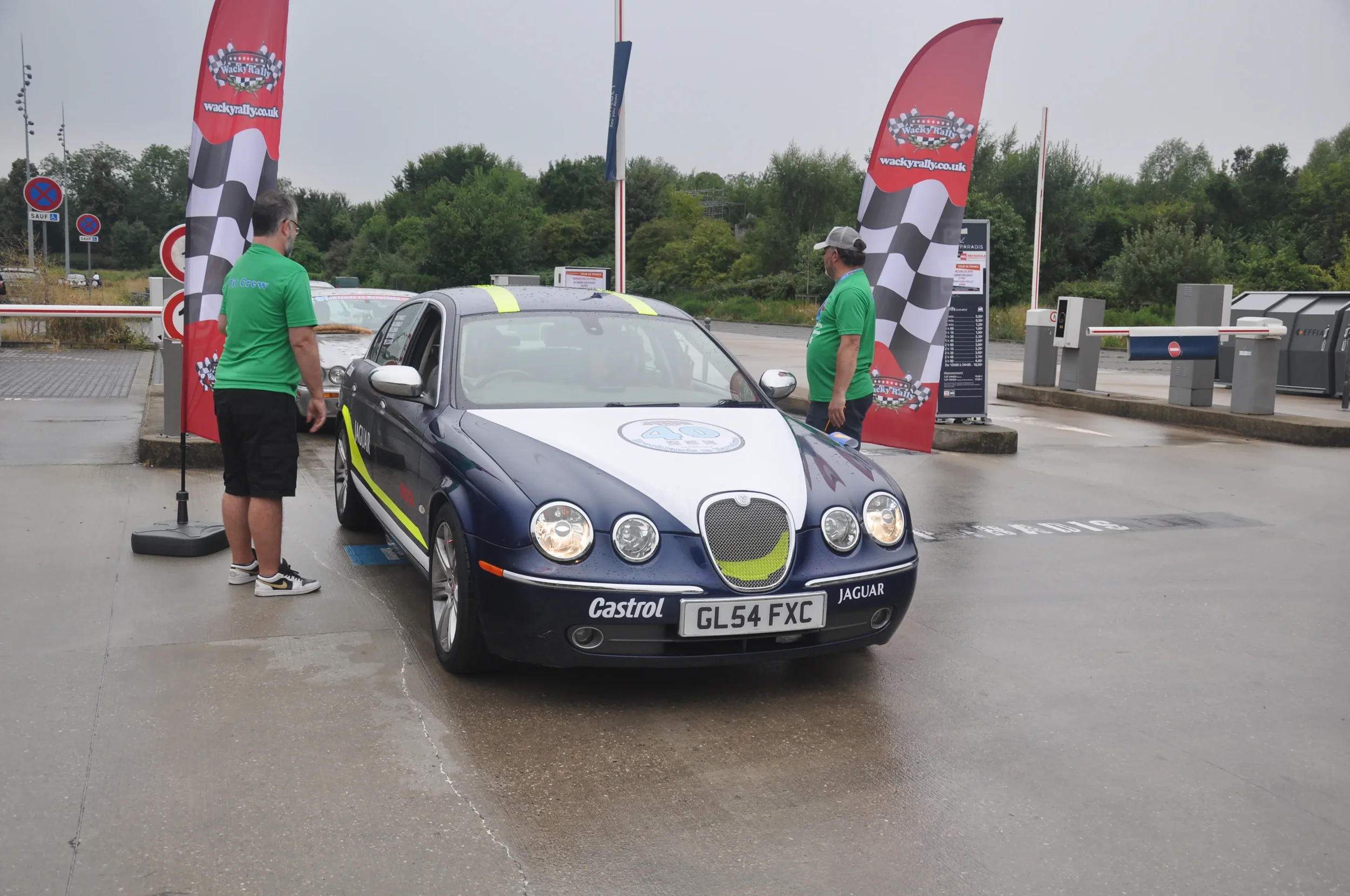 A blue Jaguar car with a white and yellow racing stripe parked at a race track with two men in green shirts standing nearby. There are red and black checkered flags and a signage board in the background.