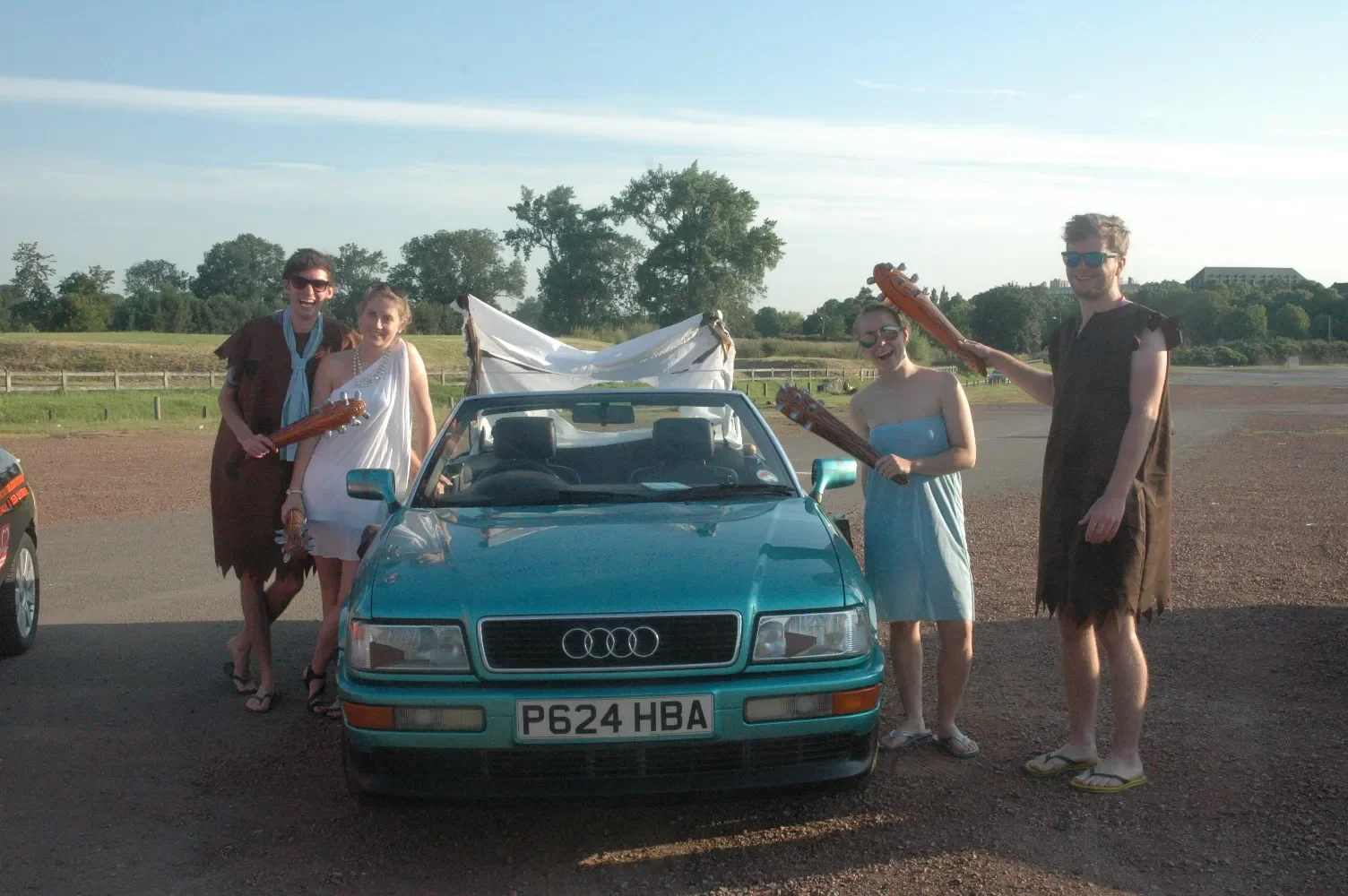Five young adults dressed as cavemen and women standing around a turquoise vintage Audi convertible with a white blanket or sheet behind it. They are holding toy axes and smiling, in an outdoor parking lot with trees and a field in the background, un