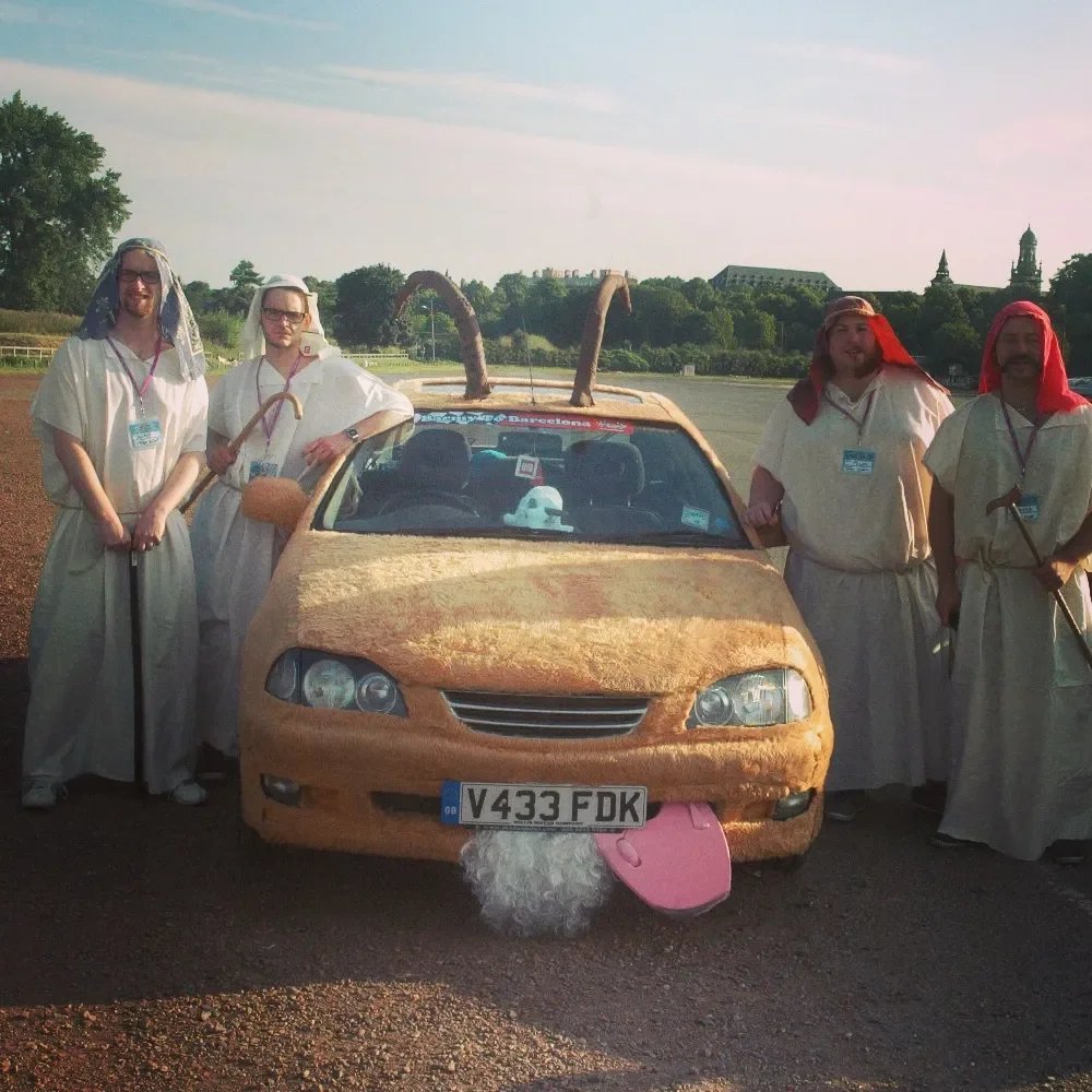 Five people dressed as biblical characters stand next to a car covered in fake dirt and mud, with a sausage-like object on the roof and a pink tongue hanging from the front bumper, resembling a camel.