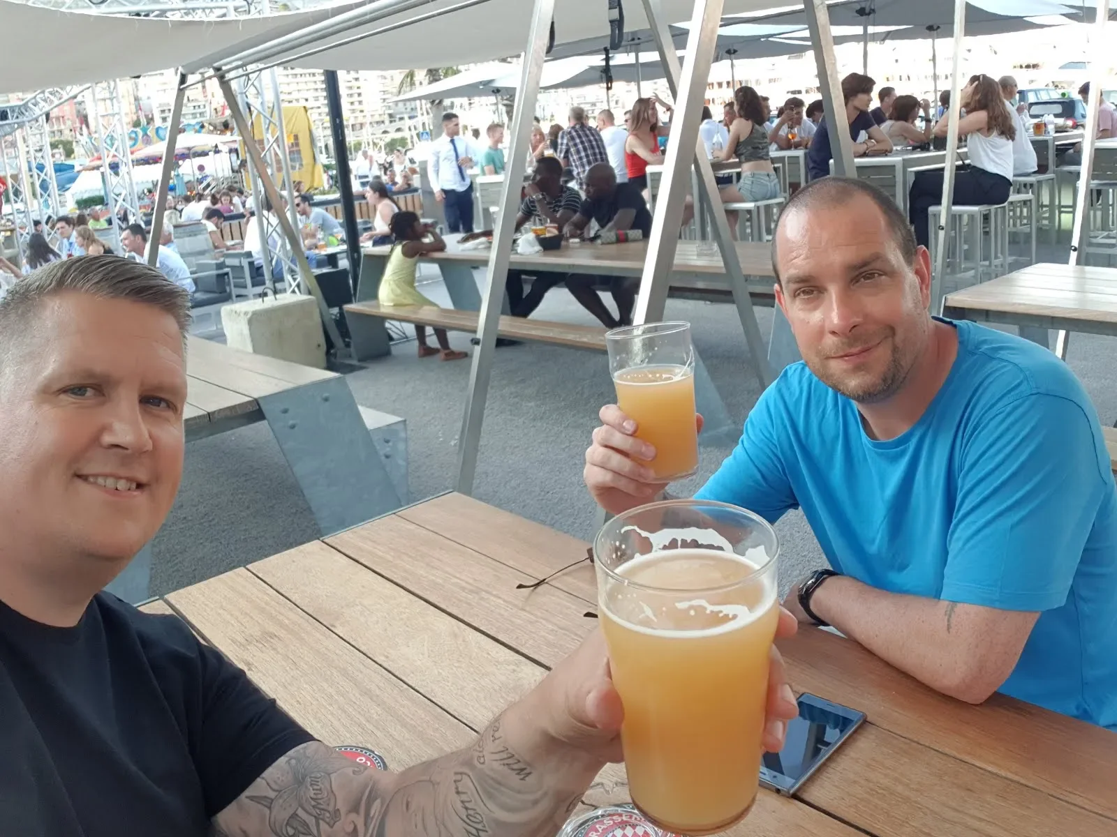 Two men sitting at a wooden table outdoors, holding glasses of pale yellow beverage, smiling, with a busy waterfront restaurant in the background.