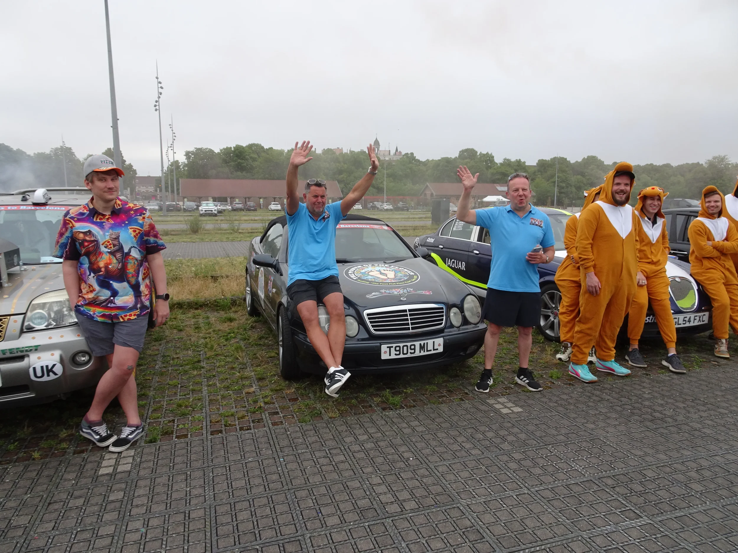 Group of people standing and sitting near parked cars, some in lion costumes, outdoors on a cloudy day