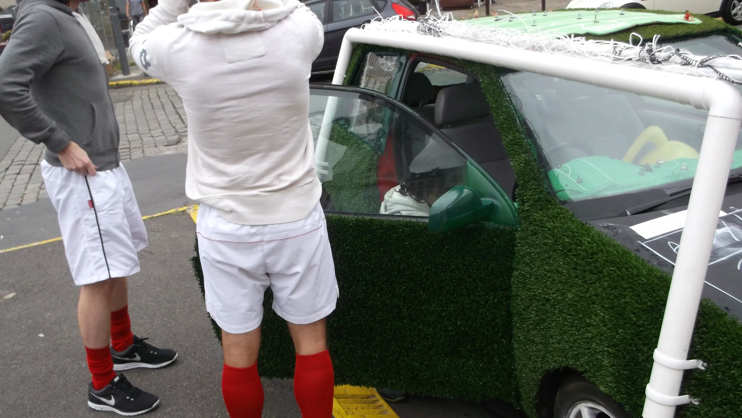 Two people standing near a small car decorated with artificial grass and a large white frame, possibly for an event or parade.