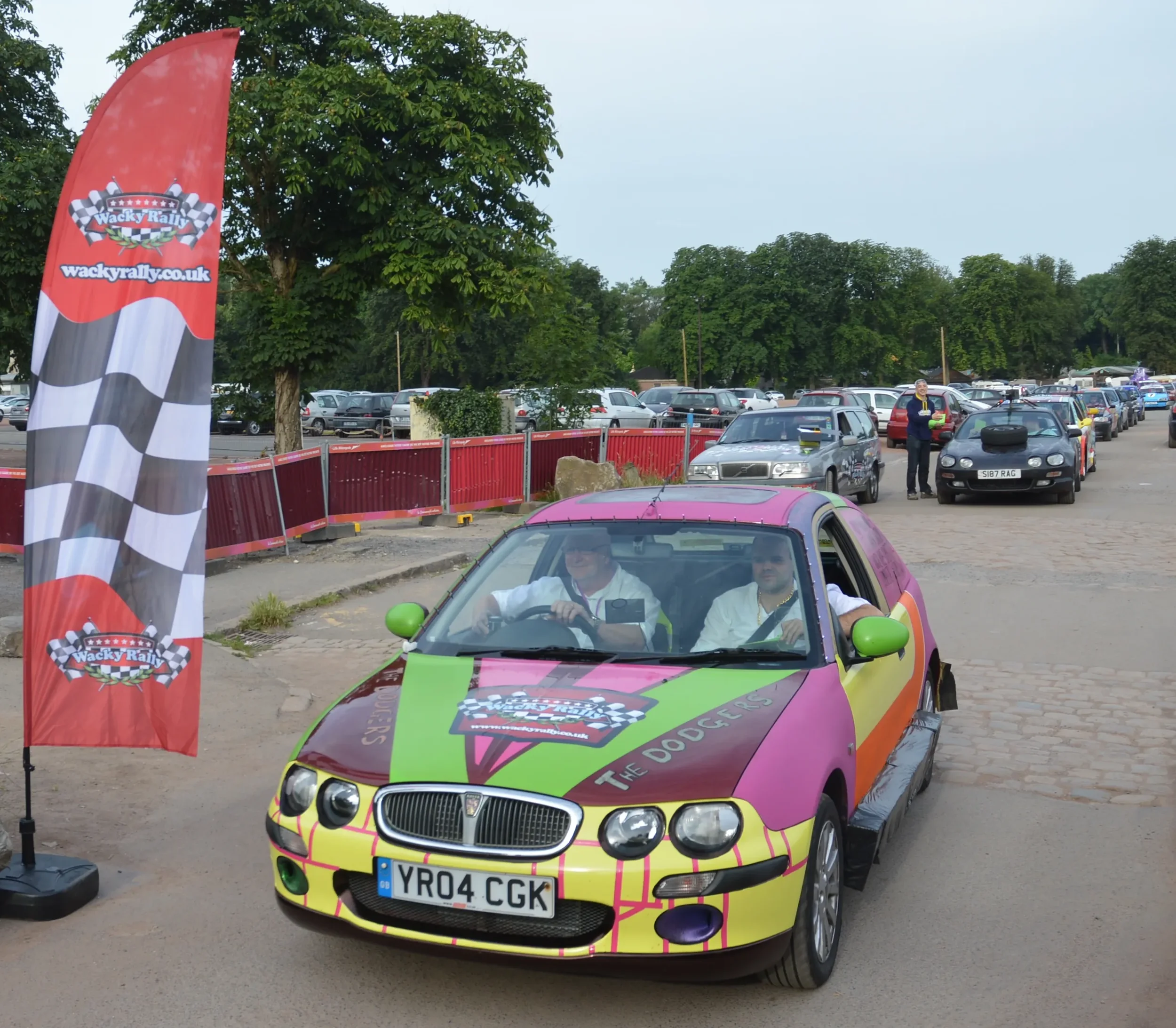 A colorful race car with a pink, green, and yellow paint job featuring the logo 'The Doggers' and a checkered flag pattern, parked on a dirt and cobblestone surface near a red and black flag with the 'Wacky Rally' logo, at a motorsport event. Two peo