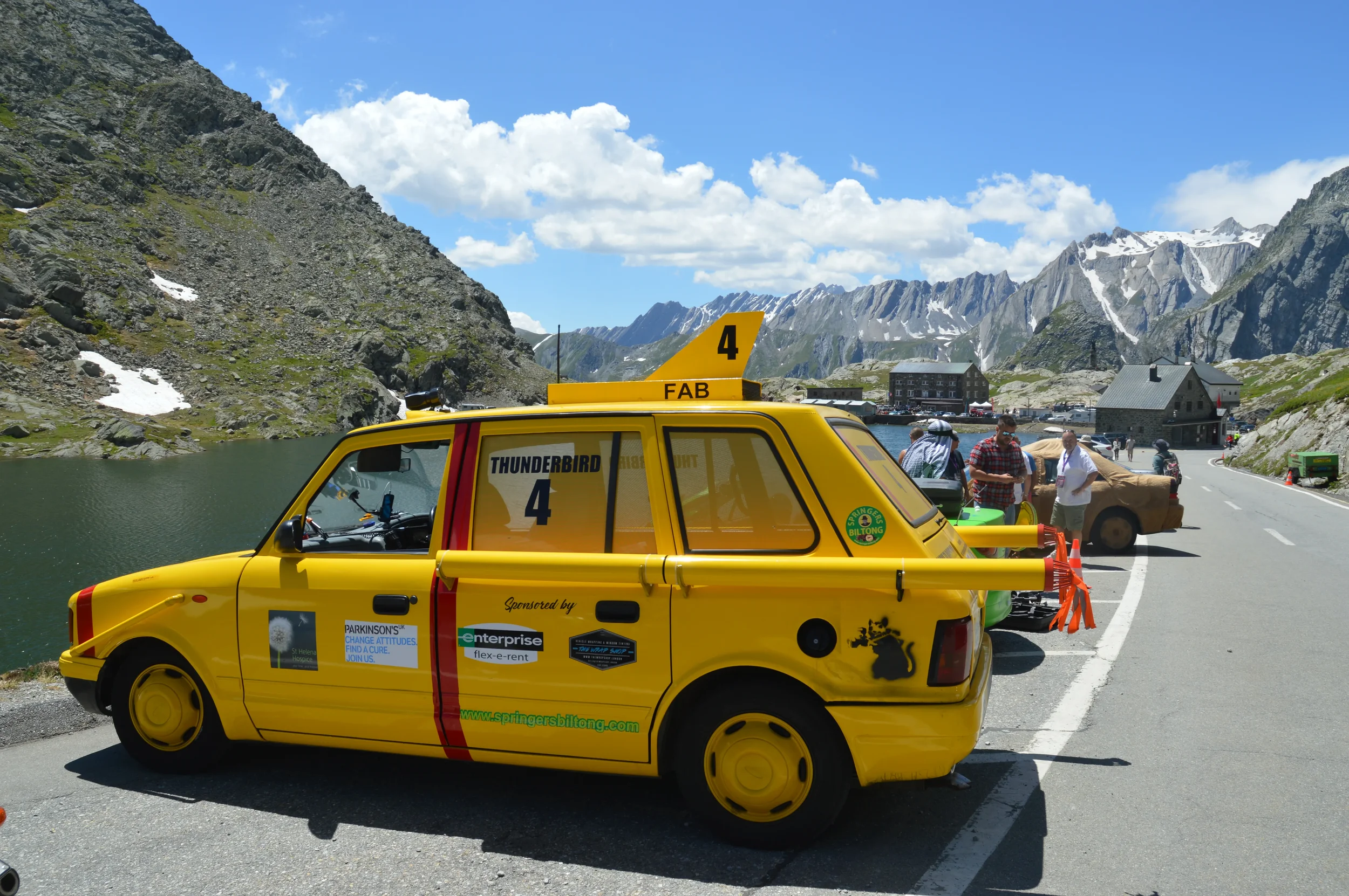 Yellow car labeled 'Thunderbird 4' parked beside a mountain lake, with mountains, clouds, and a clear blue sky in the background. Several people stand nearby, and a few cars are parked along a winding mountain road.
