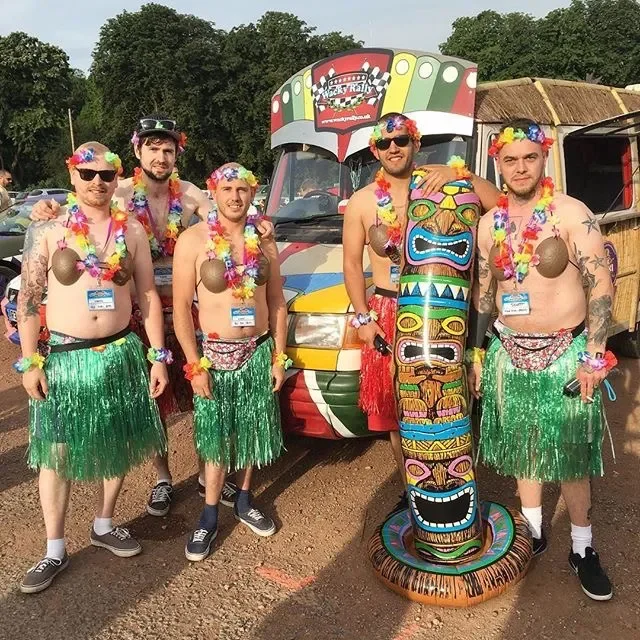 Five men dressed in tropical-themed costumes with grass skirts, leis, and floral accessories, standing in front of a colorful bus at an outdoor event. One man holds a large carved tiki statue.