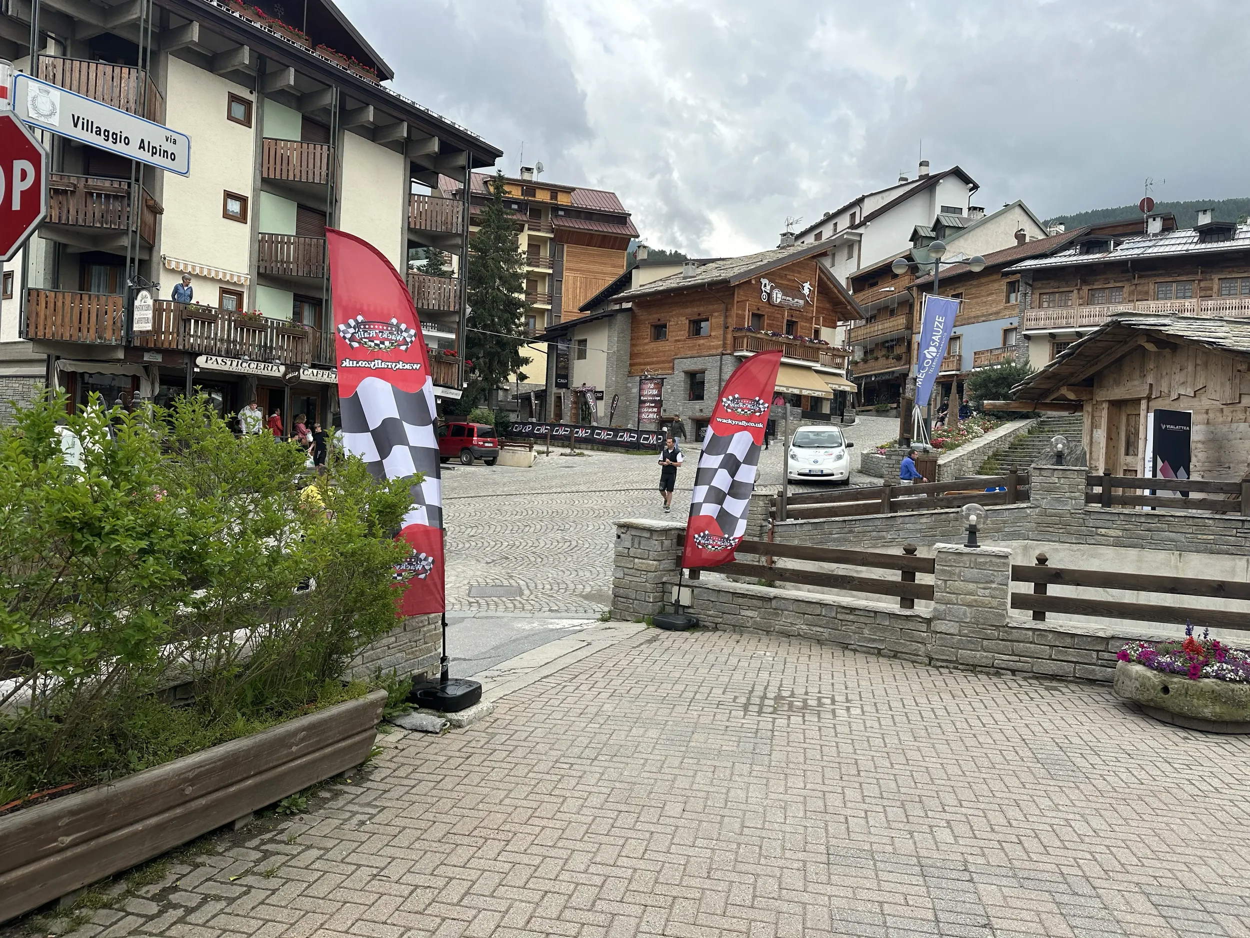 A European-style mountain village with wooden and stone buildings, colorful flags with racing checkered pattern, leather seating, and cobblestone pavement, under an overcast sky.