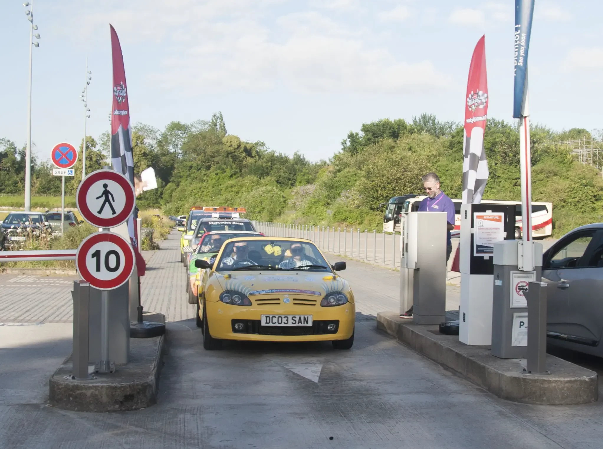 A line of colorful cars waiting at a tollbooth on a highway, with a man at the toll plaza. Signs indicate a pedestrian zone and a speed limit of 10 km/h. Flag banners are present.