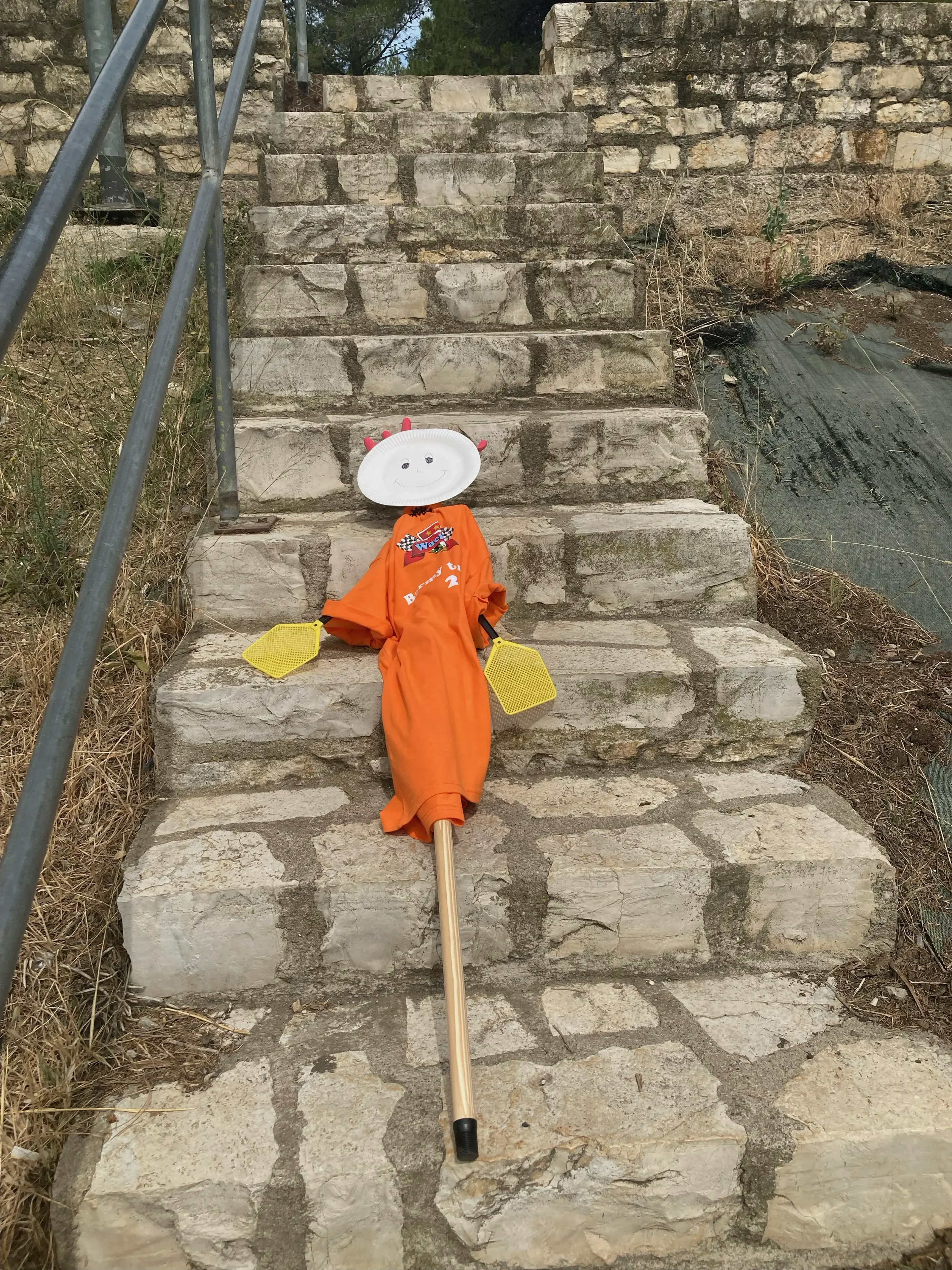 A scarecrow made from a paper plate with a smiling face, a white stick for a body, dressed in an orange shirt, positioned on stone steps. The scarecrow has red plastic spikes on the paper plate, yellow foam shapes for hands, and is leaning back again