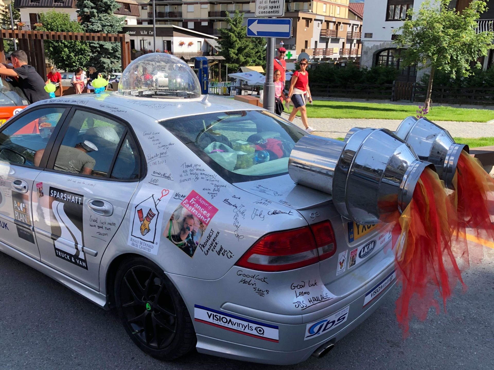 A silver race car decorated with notes, signatures, and stickers, with large exhaust pipes adorned with colorful tulle or fabric, parked on a street with people and buildings in the background.