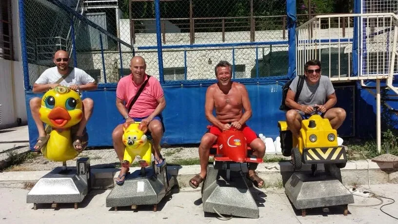 Four men sitting on amusement ride cars shaped like animals and vehicles at an outdoor amusement park.
