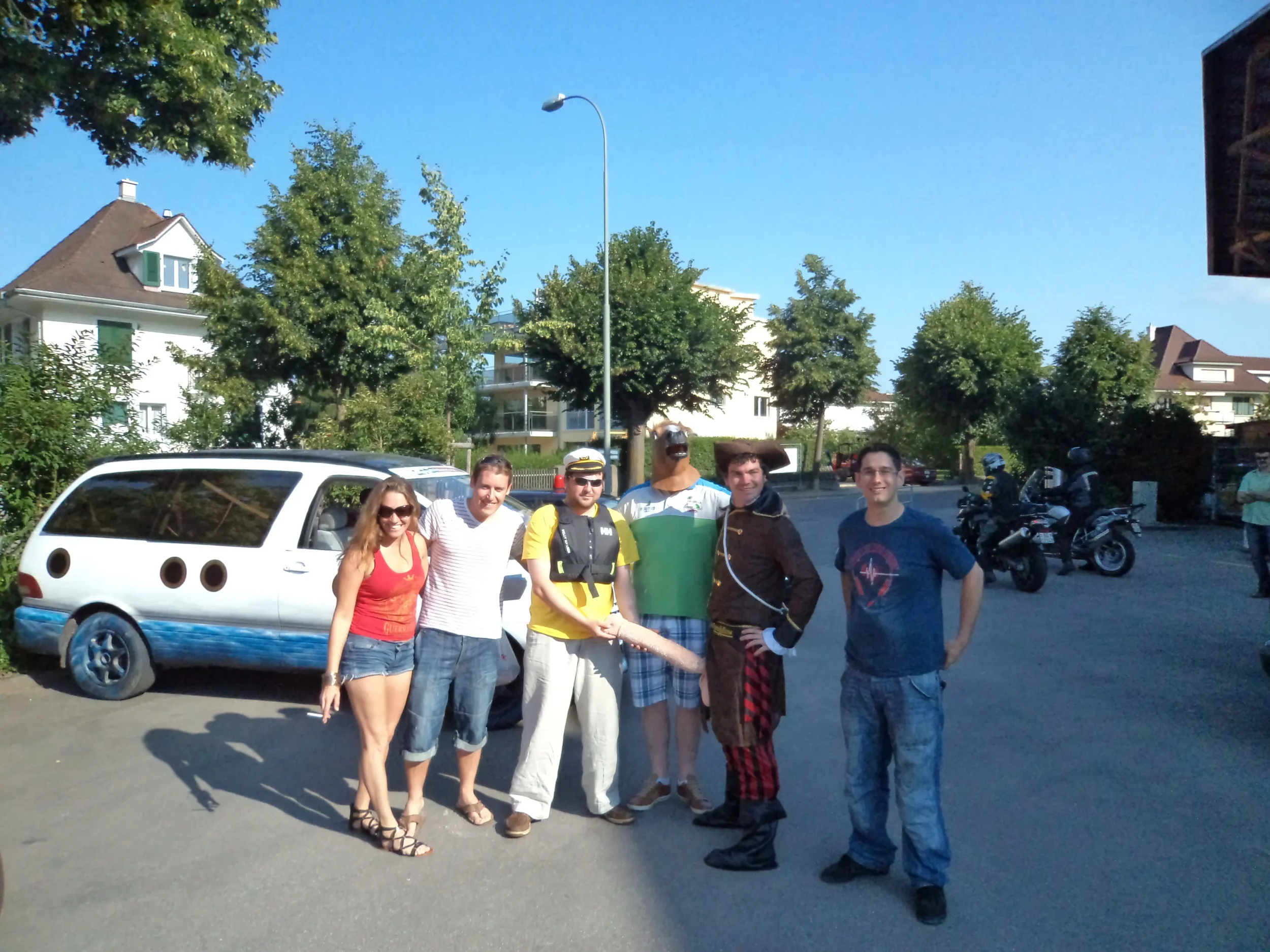 Group of six people standing outdoors on a sunny day, with trees, houses, a white van, and motorcycles in the background.