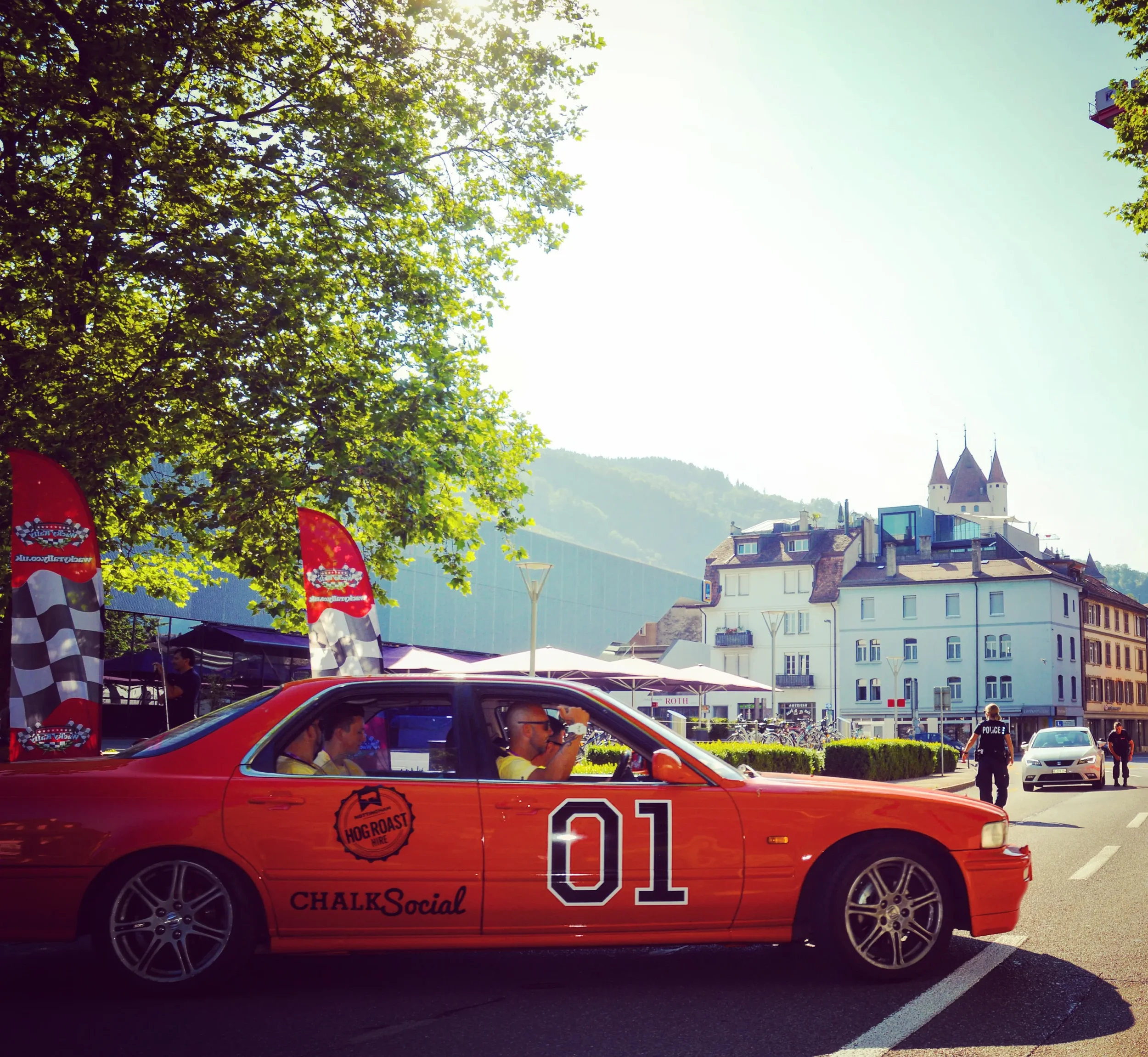 Red sports car with the number 01 and CHALKSocial and Hog Roast logos parked on a street with trees, buildings, and a police officer walking nearby.
