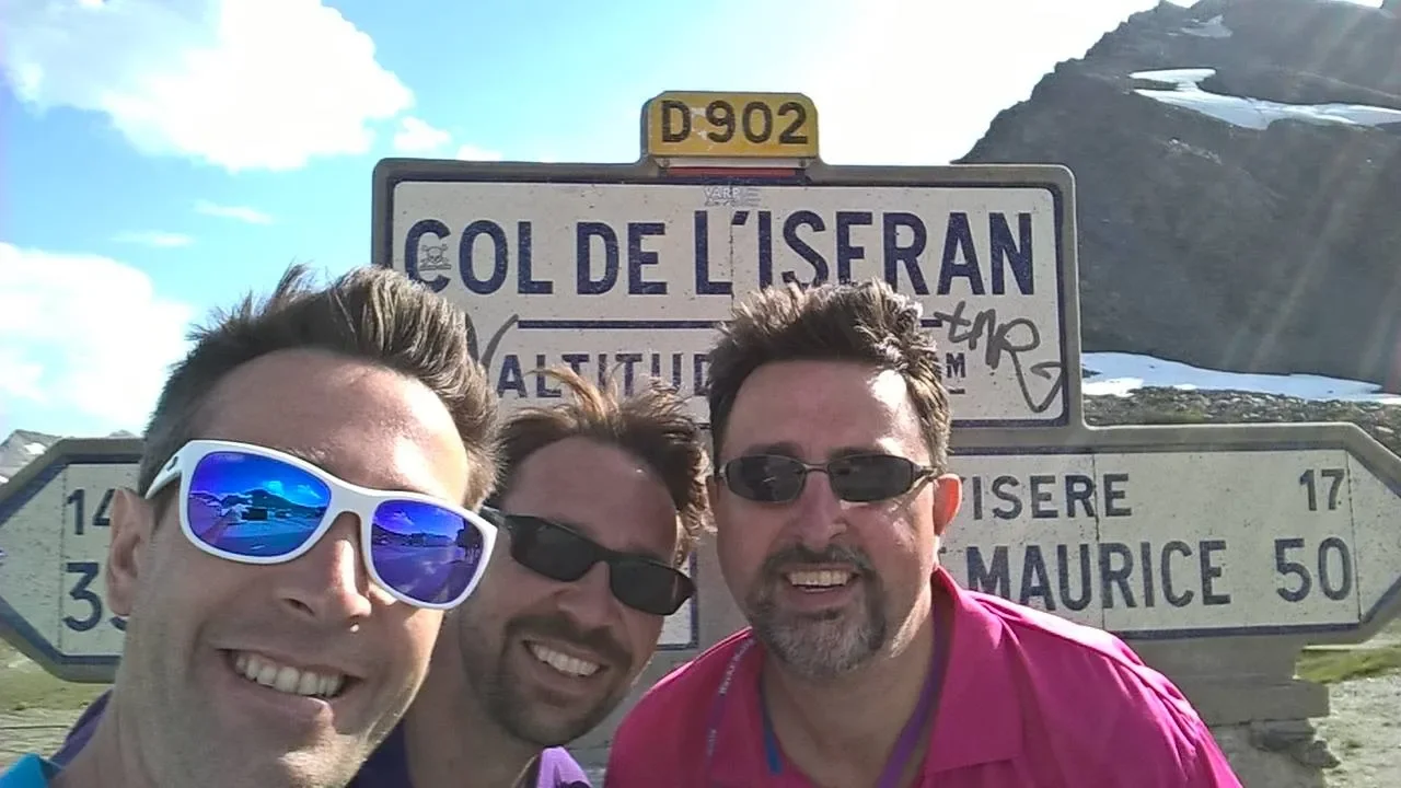 Three men smiling with sunglasses taking a selfie in front of a mountain sign that reads 'Col de l'Iseran' in France.
