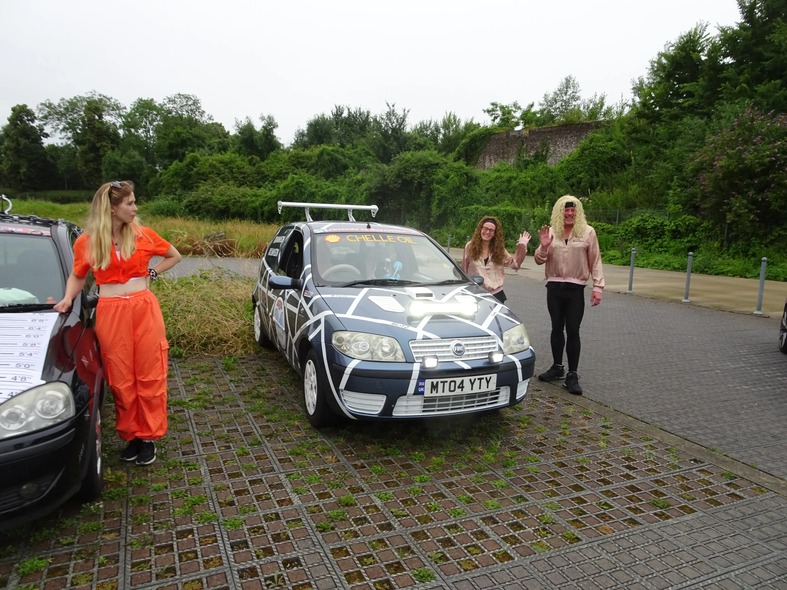 Four people standing near a blue and white rally car with 'Chell Oil' on the windshield, some waving, in an outdoor parking lot with greenery in the background.
