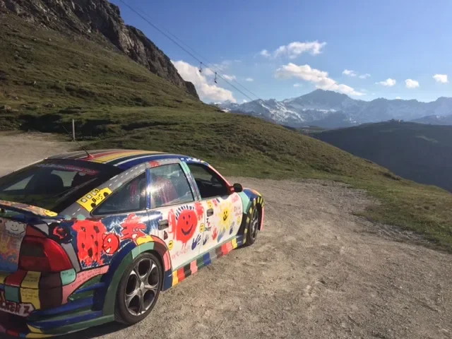 A colorful car with various cartoon and graffiti-style artwork parked on a dirt area in a mountainous landscape with green hills, rocky cliffs, and snow-capped peaks in the background under a partly cloudy sky.