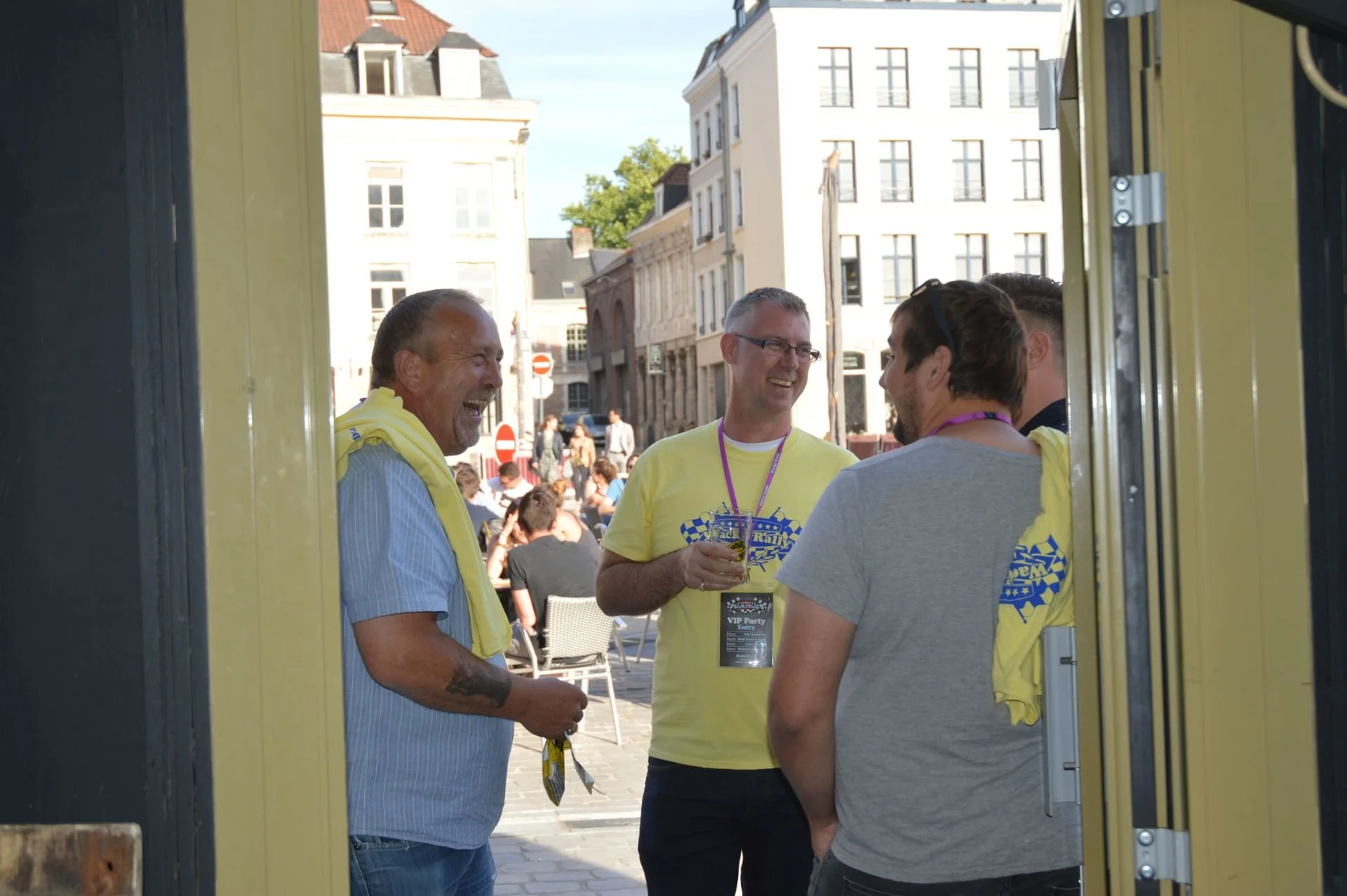 Four men are standing and talking outside, some wearing yellow shirts and VIP lanyards, in a city square with people sitting at tables and historic buildings in the background, during daytime.