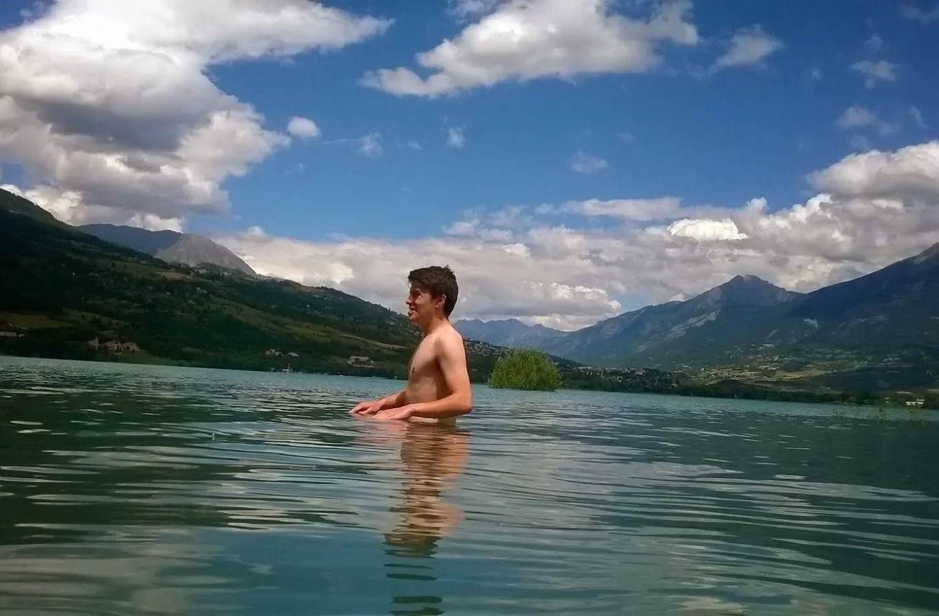 A shirtless young man standing in a lake with mountains in the background under a partly cloudy sky.