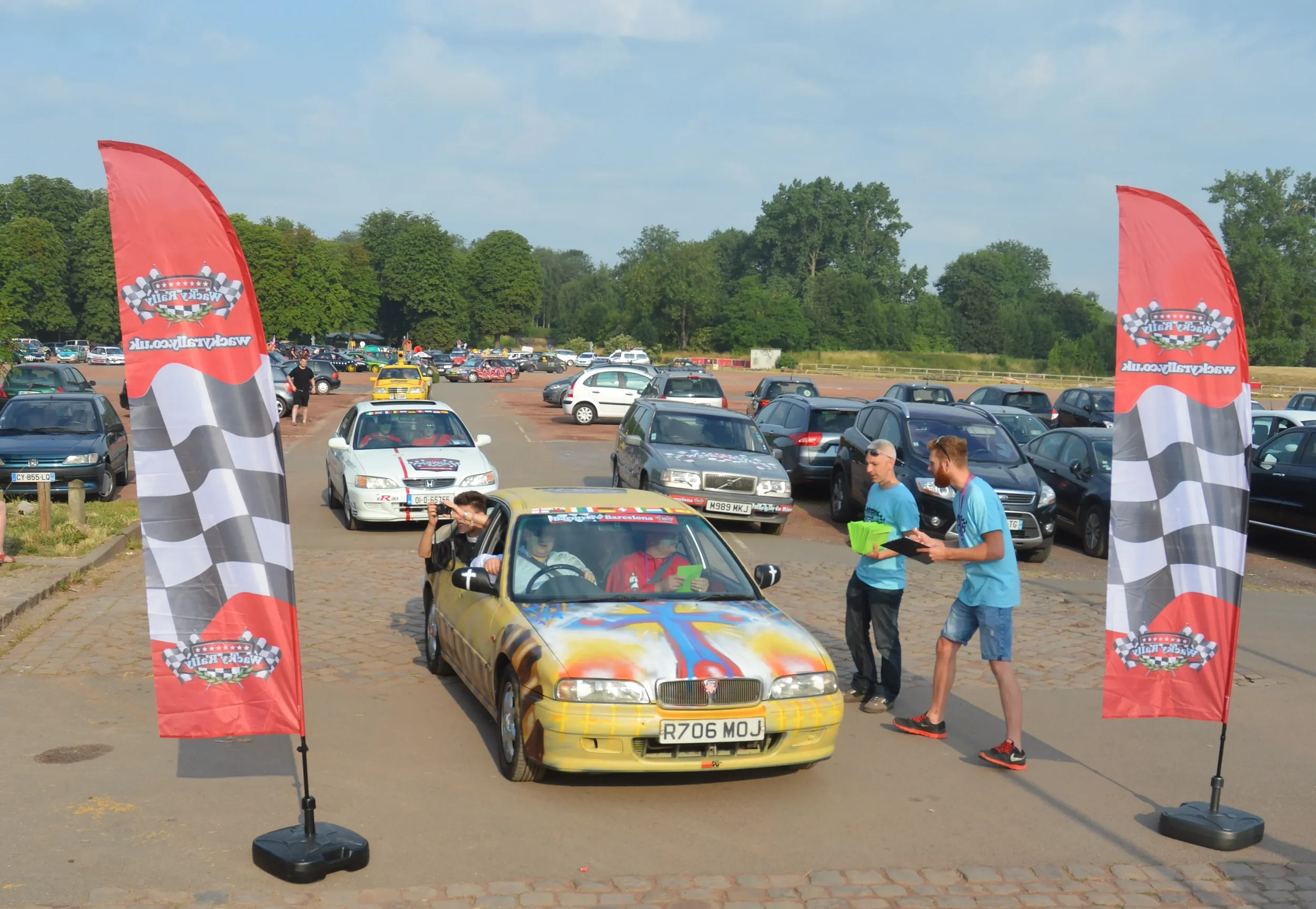 A colorful, painted car at a racing event, with two people standing beside it holding clipboards, and another person sitting inside the car. The scene is set in a parking lot filled with cars, with checkered flags on either side and a clear, sunny sk