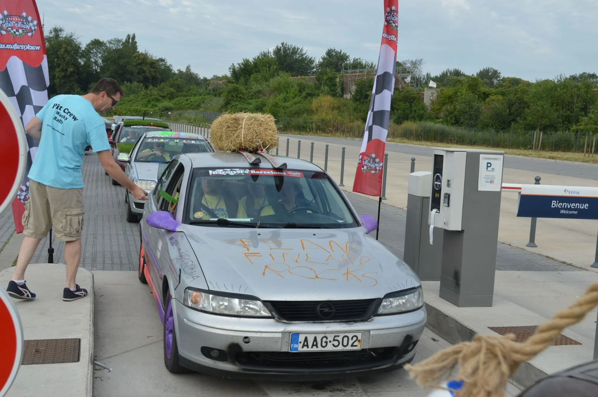 A line of parked cars at a racetrack, with a man in a blue T-shirt checking a silver car with a large hay bale on the roof. The car has a message written on the hood with a windshield banner reading 'Ready to Barcelona' and a license plate 'AAG-502.'