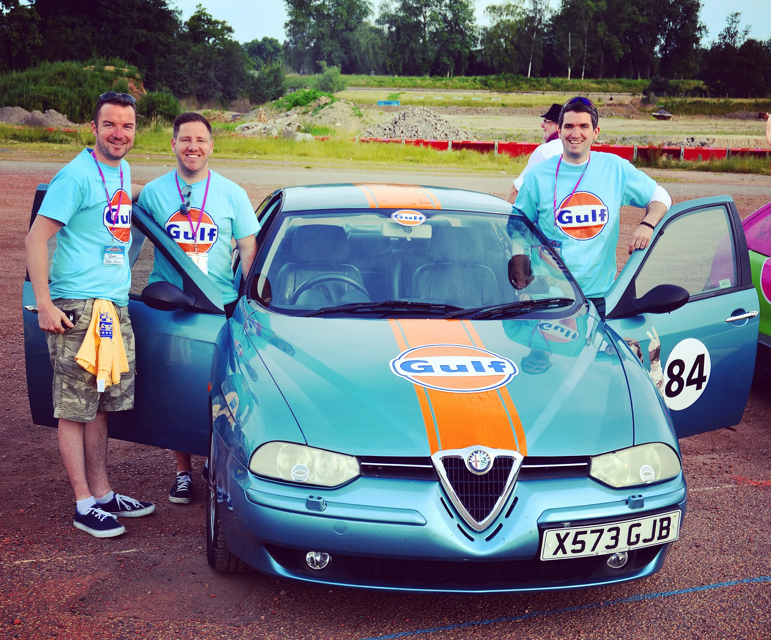 Four men standing beside a blue Alfa Romeo race car with Gulf logo, on a dirt racing track, with greenery and trees in the background, wearing Gulf T-shirts and smiling.