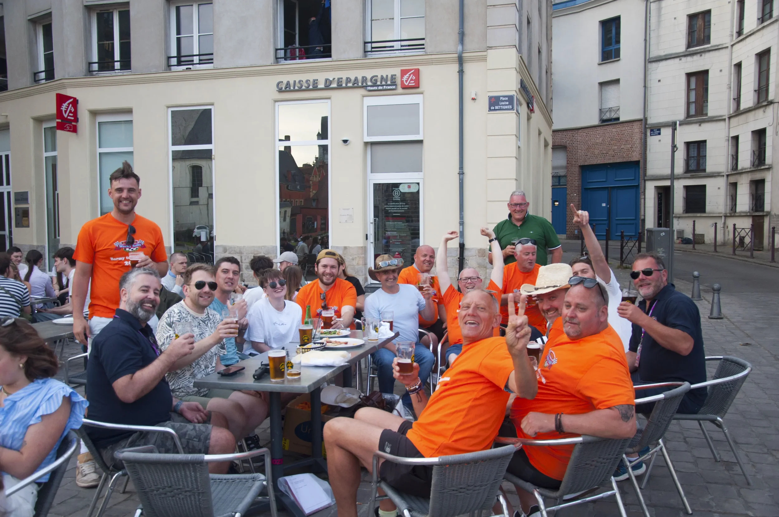 Group of people sitting at an outdoor cafe with drinks, smiling, raising glasses, and enjoying a sunny day in front of a building with a sign that says "Caisse d'Epargne."
