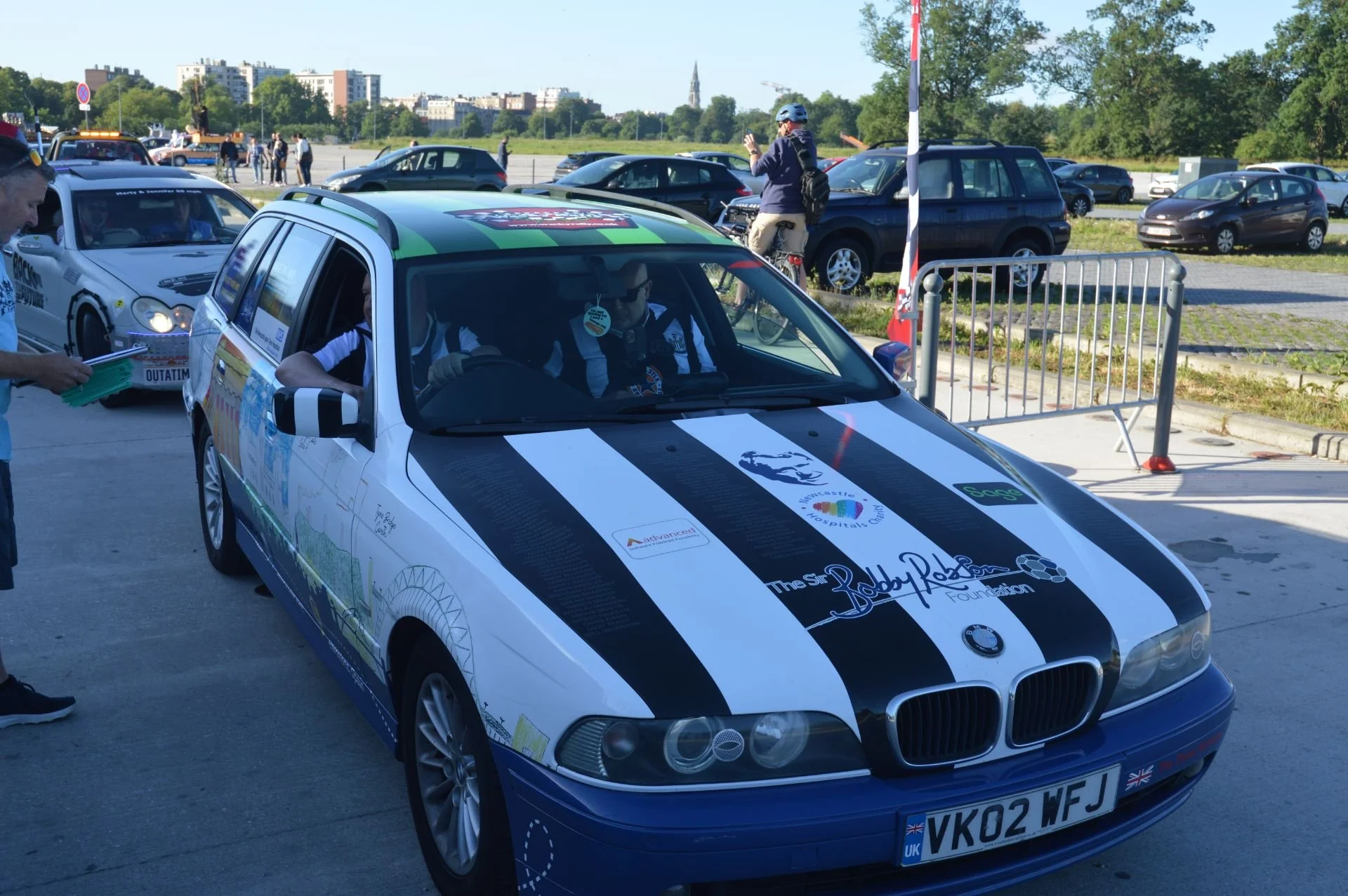 A white BMW race car with black and white racing stripes and custom decals, including 'The Robby Rob Foundation,' parked outdoors with a man inside wearing sunglasses. In the background, there are people, parked cars, green trees, and a cityscape.
