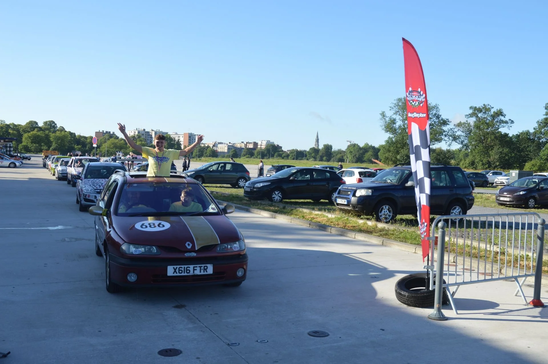Young man standing through the sunroof of a red unhealthy-looking Renault car celebrating at a racing event, with a line of parked cars and a checkered flag banner on the right side.