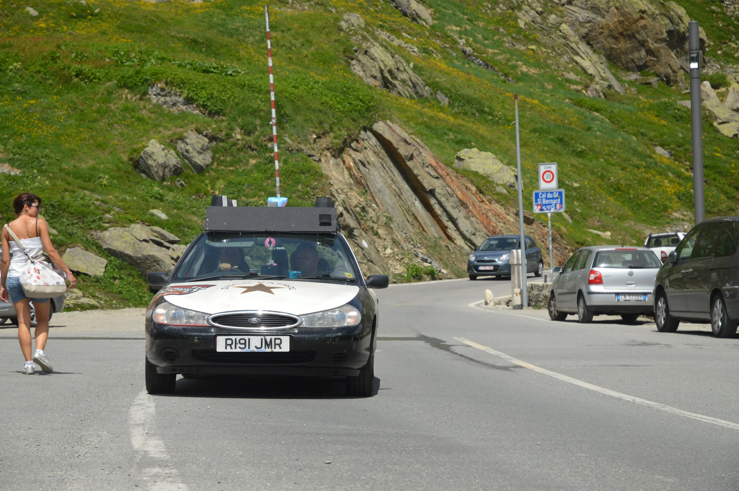 Tourist car parked on the side of a mountain road near a rock face, with a woman walking by carrying a large bag.