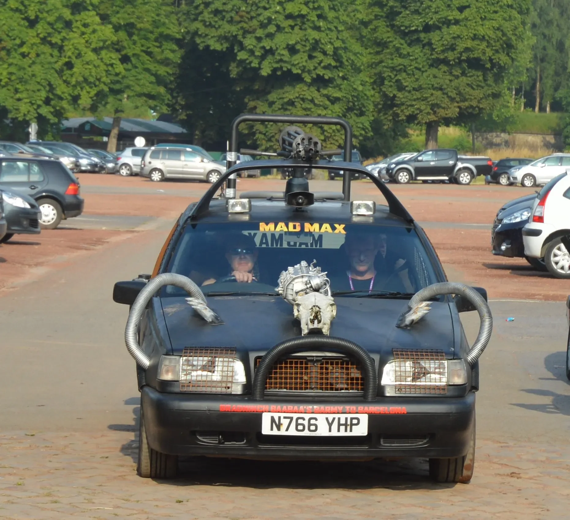 Modified car with a skull on the hood, large hoses, and various decorations, parked in a lot with other vehicles and green trees in the background.