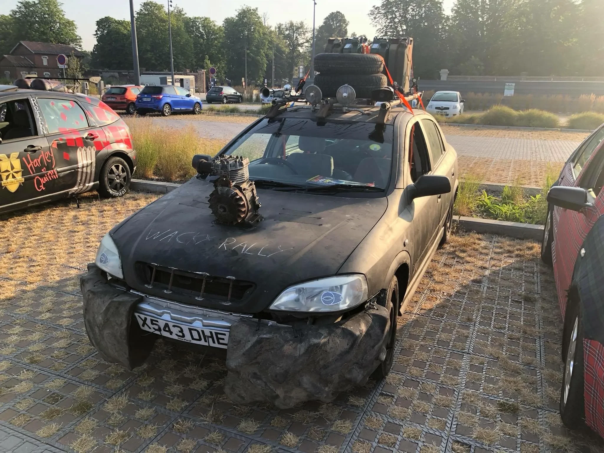 A modified car with the words 'WACKY RALLY' written on the hood, featuring a large engine mounted on the front, sturdy mud tires, and a roof rack with spare tires and equipment, parked in an outdoor lot during sunset.