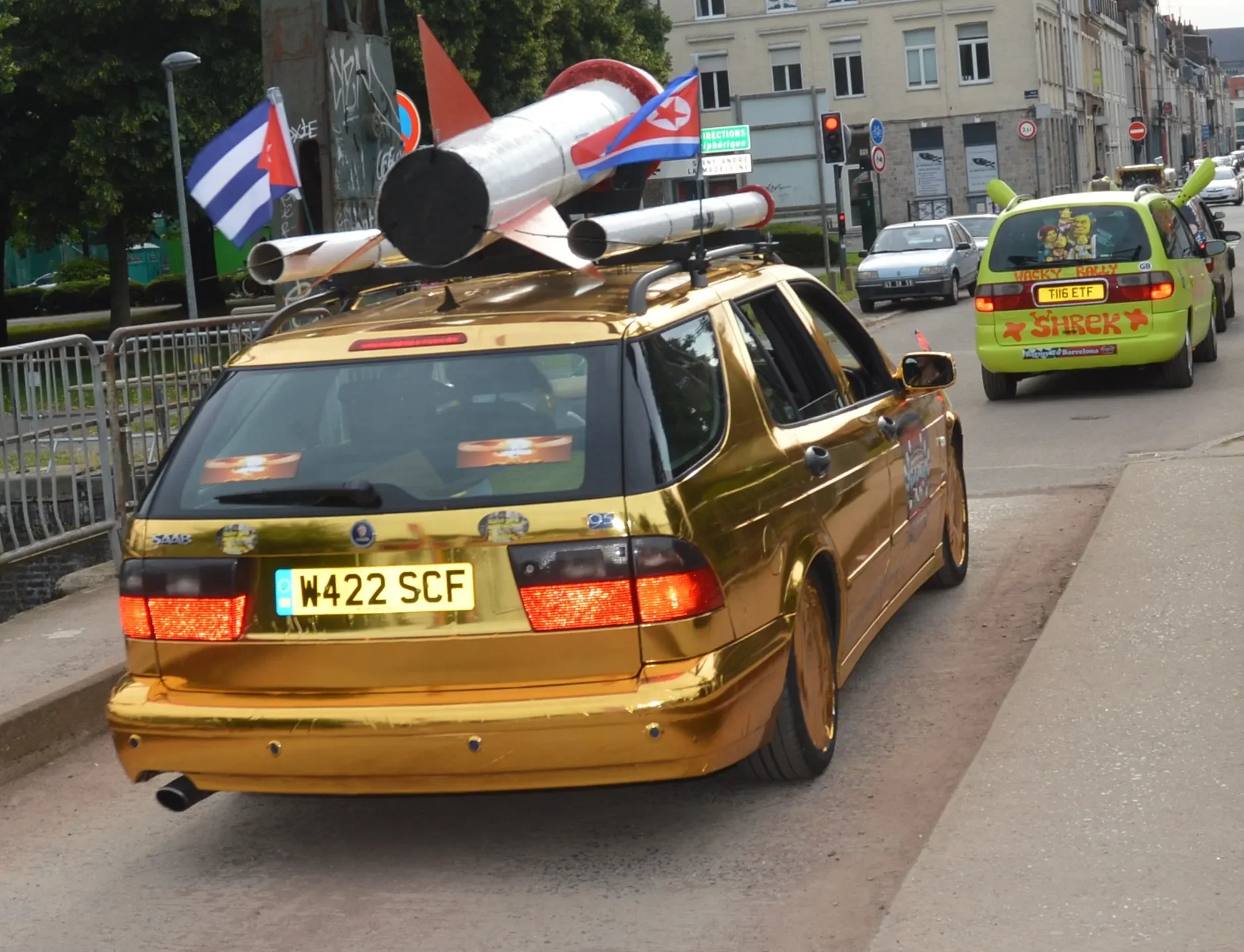A parade with a gold car decorated with stickers, with a missile replica on its roof, followed by a green vehicle with cartoon stickers and a sticker that says "Shrek."