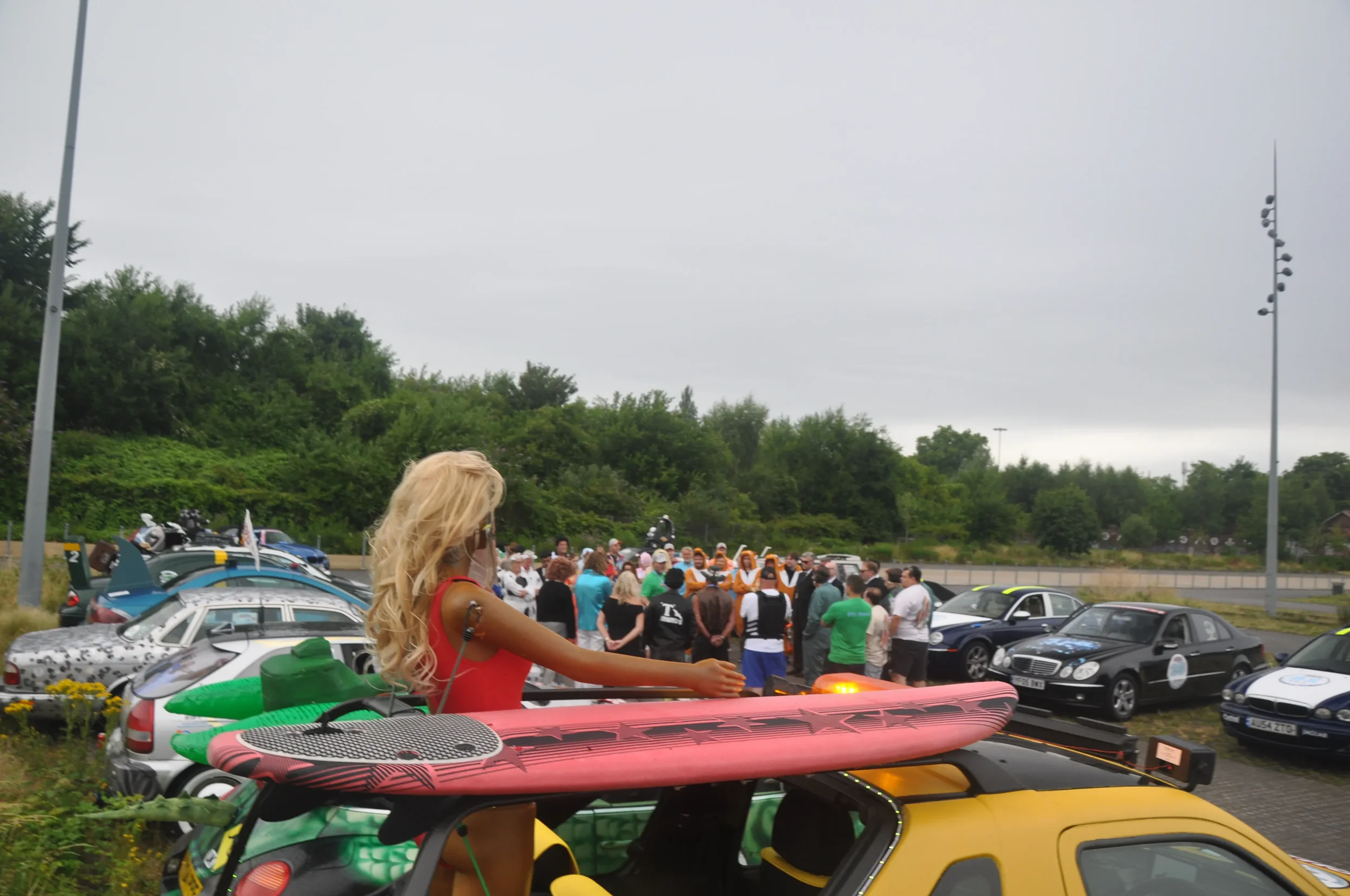 A woman with long blonde hair standing on a yellow vehicle with a pink paddleboard and green kayak on top, overlooking a crowd of people gathered near a row of parked cars on an overcast day.