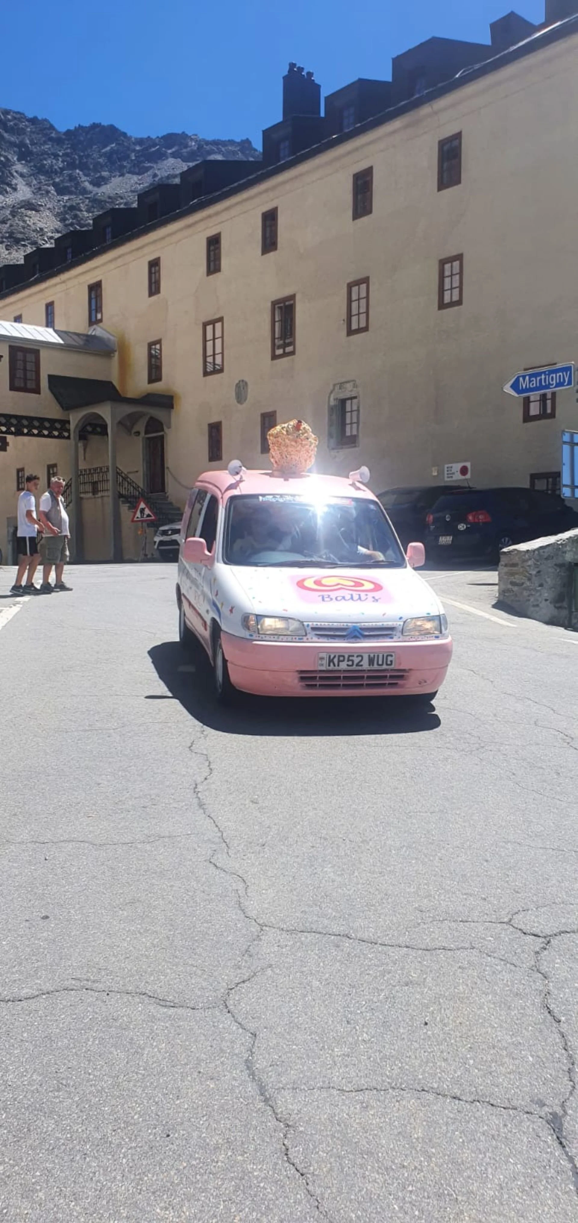 A small pink ice cream truck on a street in a mountainous area, with a large ice cream cone on its roof, and a building in the background with a blue road sign pointing to Martigny. Three people are walking by on the sidewalk.