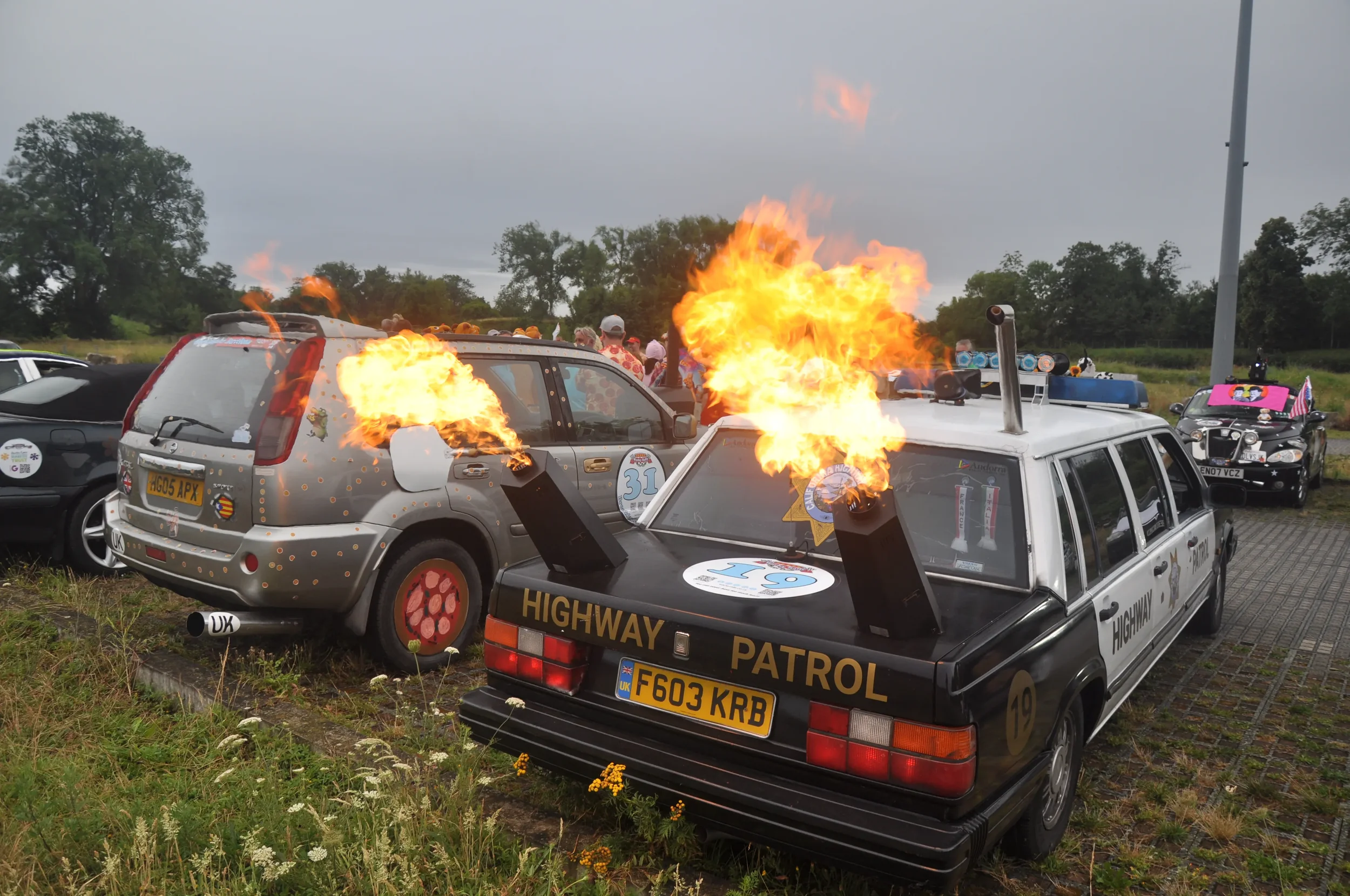 A roadside display with cars, one labeled 'Highway Patrol,' on fire with flames shooting from their hoods, part of a stunt show with spectators in the background.