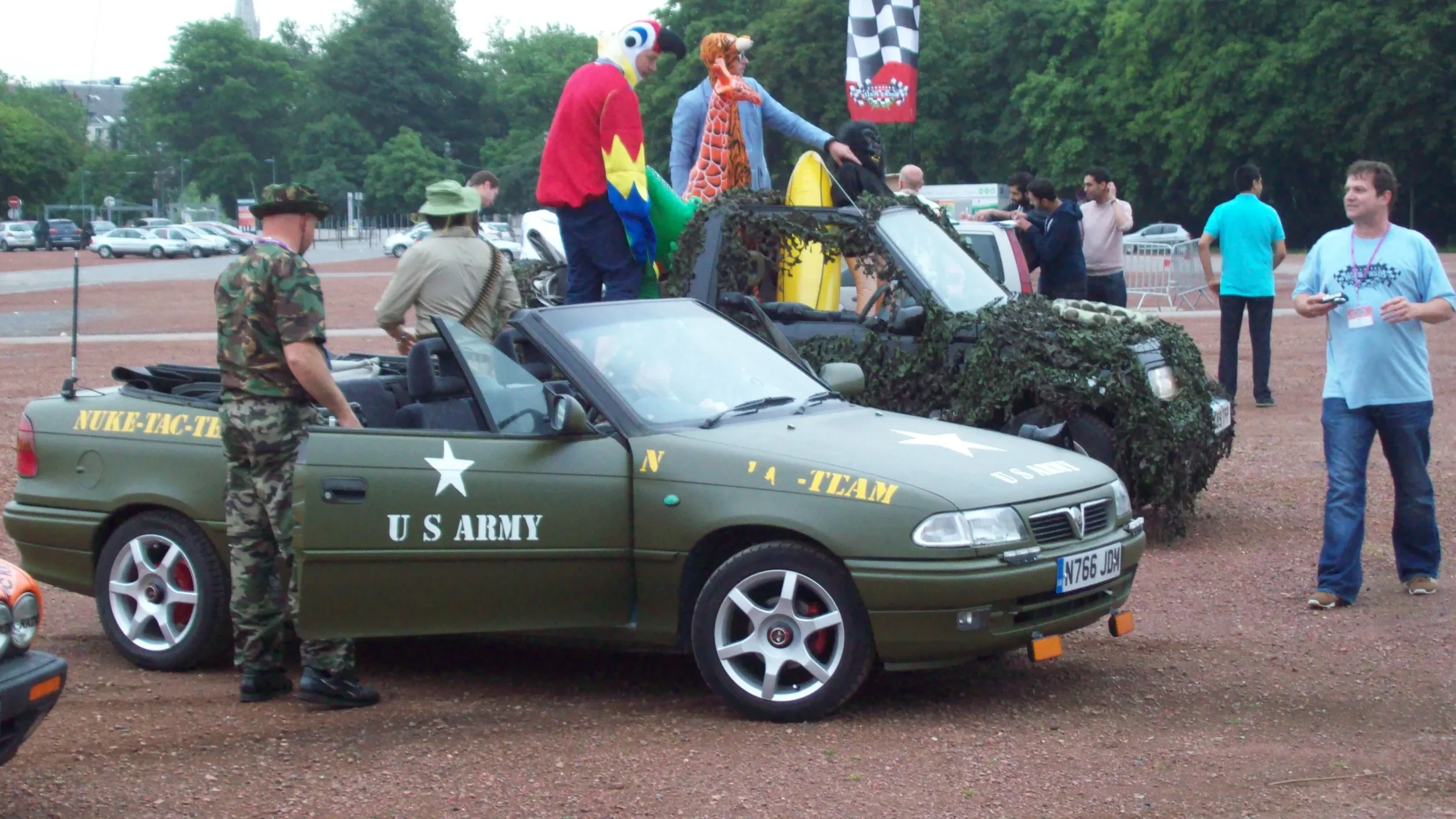 A military-themed car show featuring a green convertible with US Army markings and a camouflage SUV decorated with artificial foliage and cartoon character costumes on top. Several people are gathered around, including a man in camouflage attire and 